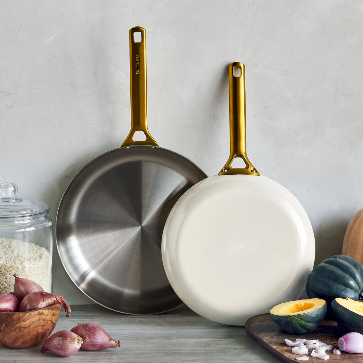 Two uncoated stainless steel frypans in cream, 10″ and 12″ sizes, on a counter with rice, onions, and vegetables nearby.
