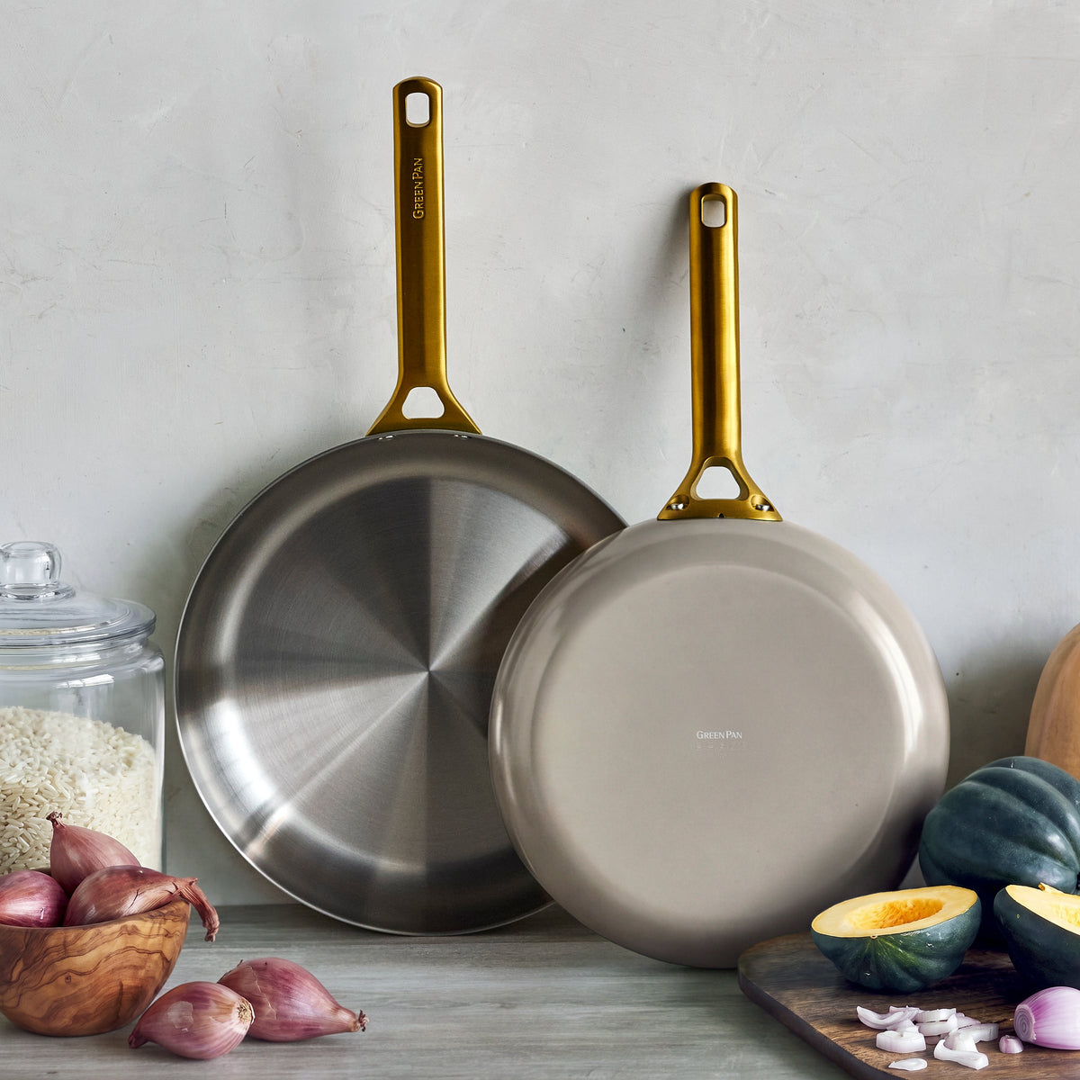 Two macadamia-colored frypans with gold handles on a kitchen counter near assorted vegetables and a jar of rice.