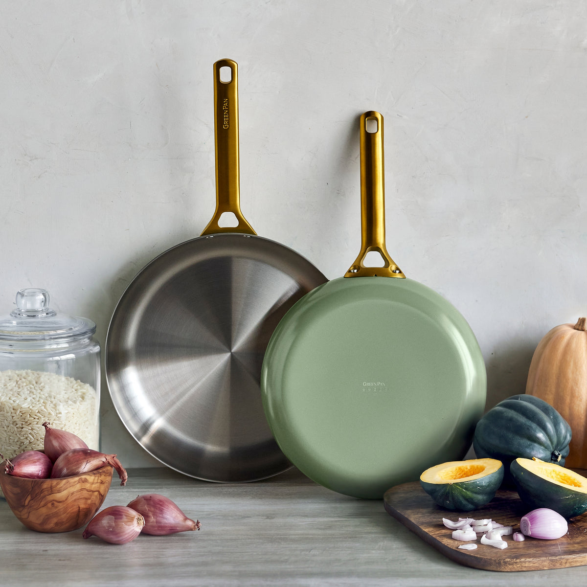 Two uncoated stainless steel frypans in sage, 10″ and 12″, on a counter with assorted vegetables nearby.