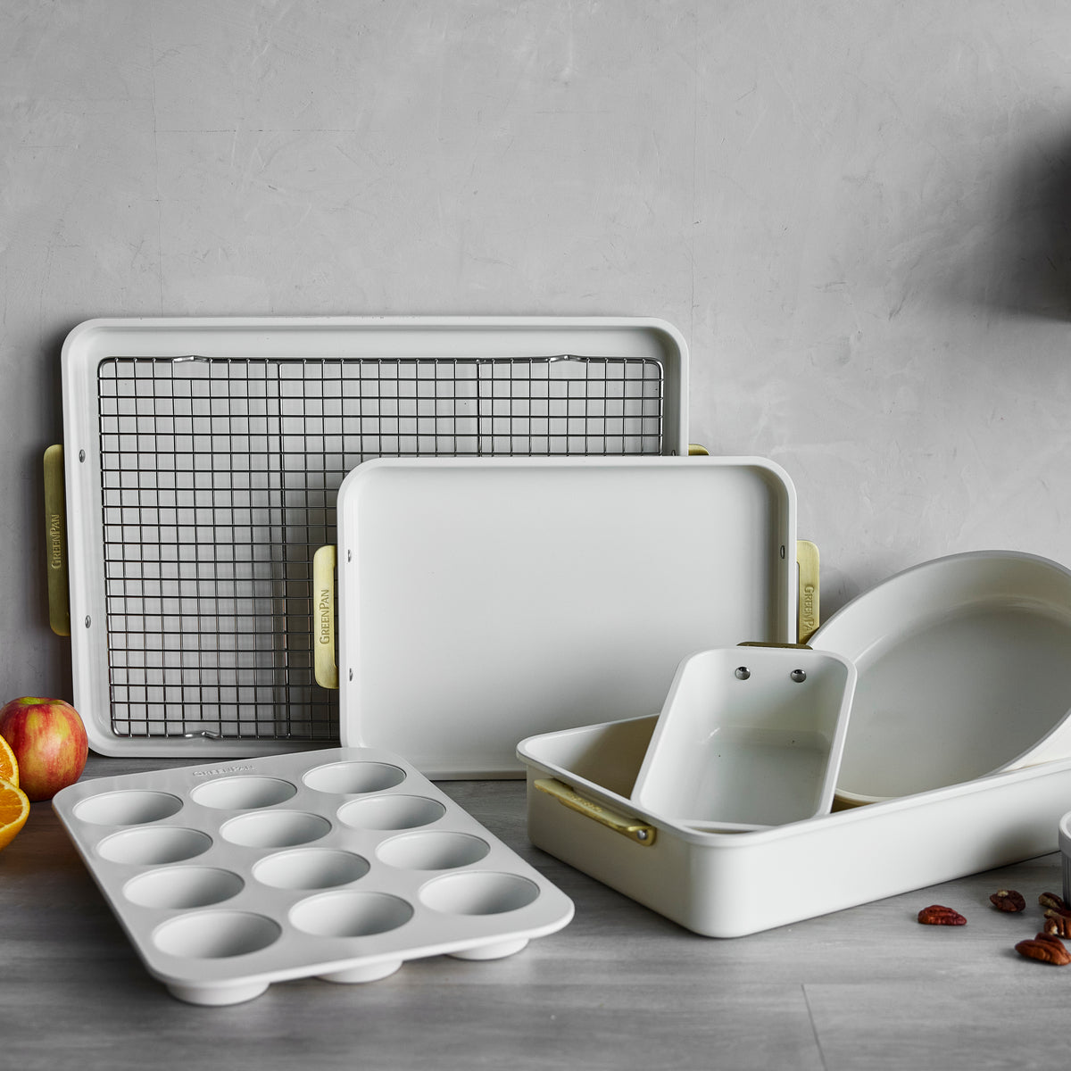 Cream and gray 7-piece bakeware set with pans, muffin tin, cooling rack, accented by fruit and nuts.