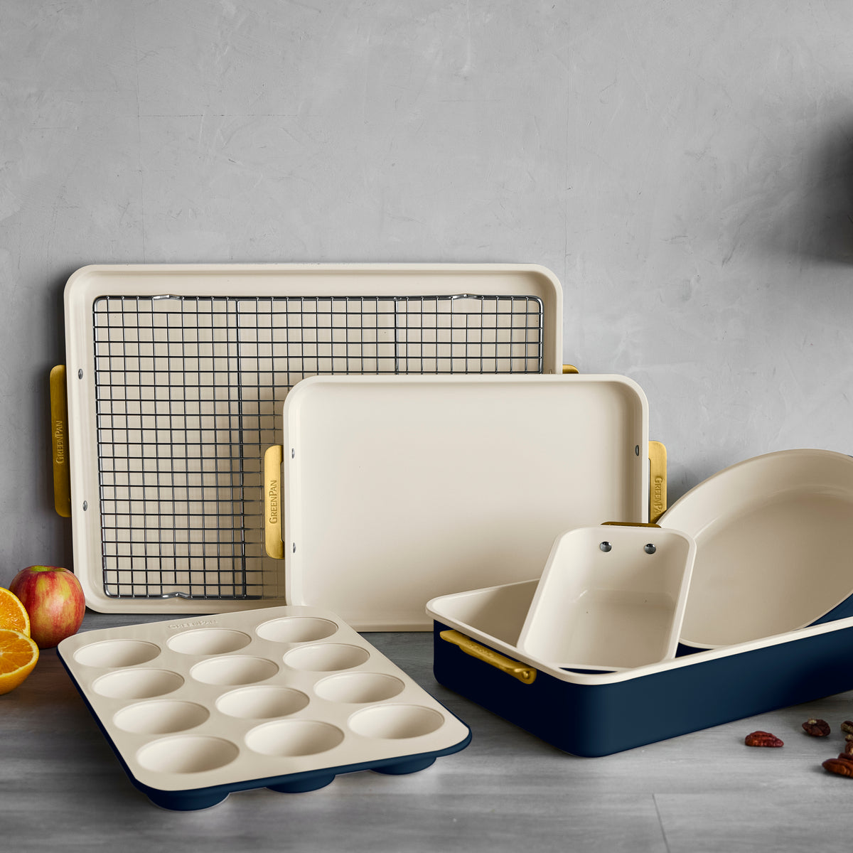 7-piece bakeware set with cream and navy trays, muffin tin, rack, spatula, and gold handles on a gray background.