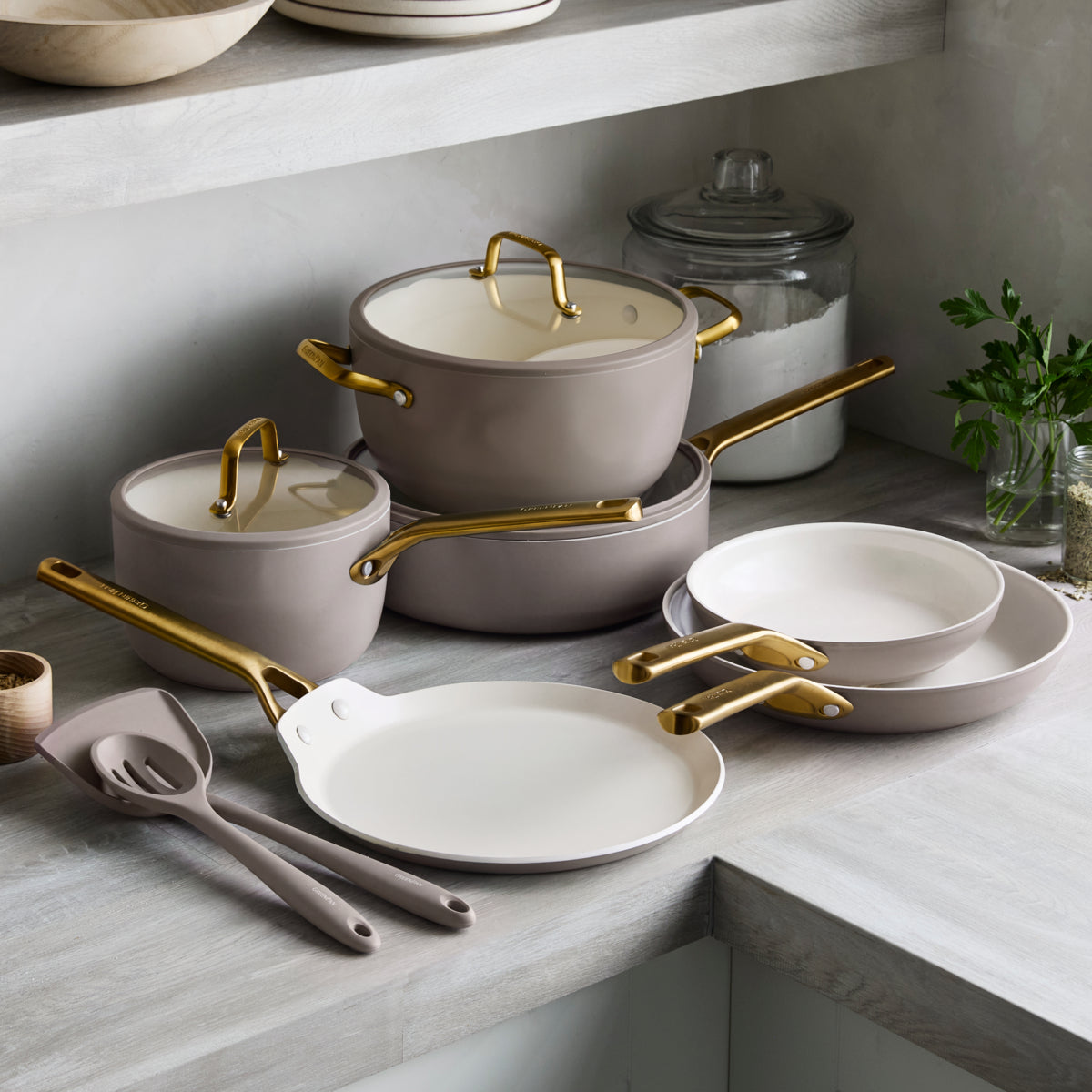 Ceramic nonstick 11-piece cookware set in latte with gold handles arranged on a kitchen counter beside utensils.