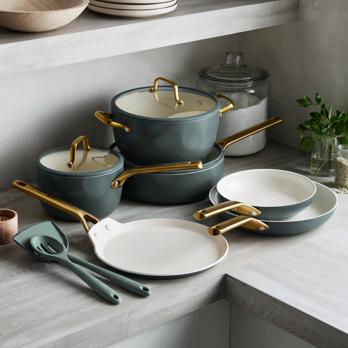 11-piece ceramic nonstick cookware set in spruce with gold handles on a counter, surrounded by fresh herbs and glass jars.