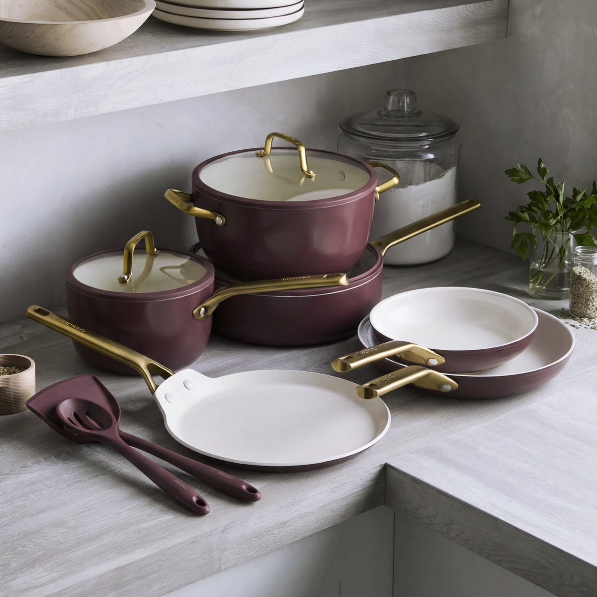 11-piece Bordeaux ceramic nonstick cookware set with gold handles on a gray counter beside herbs and a jar.