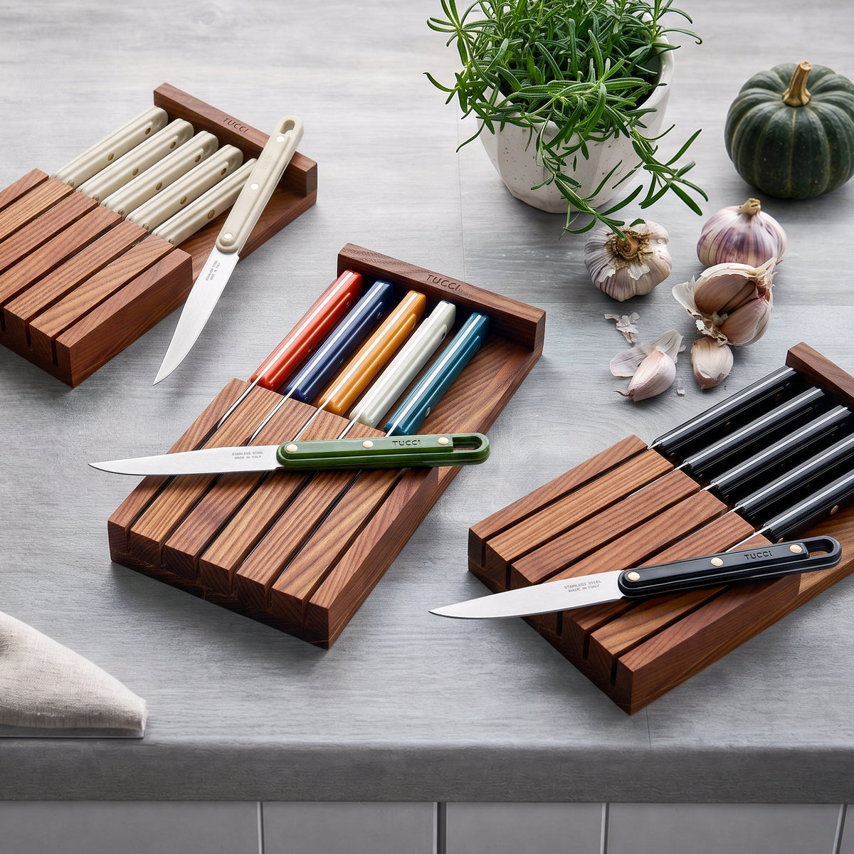 Six-piece multicolor steak knife set in a wooden tray on a kitchen counter with herbs, garlic, and a pumpkin nearby.