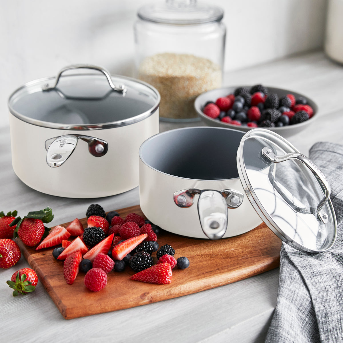 Cream ceramic 1.5-qt and 3-qt saucepans with lids on a wooden board, both opened.