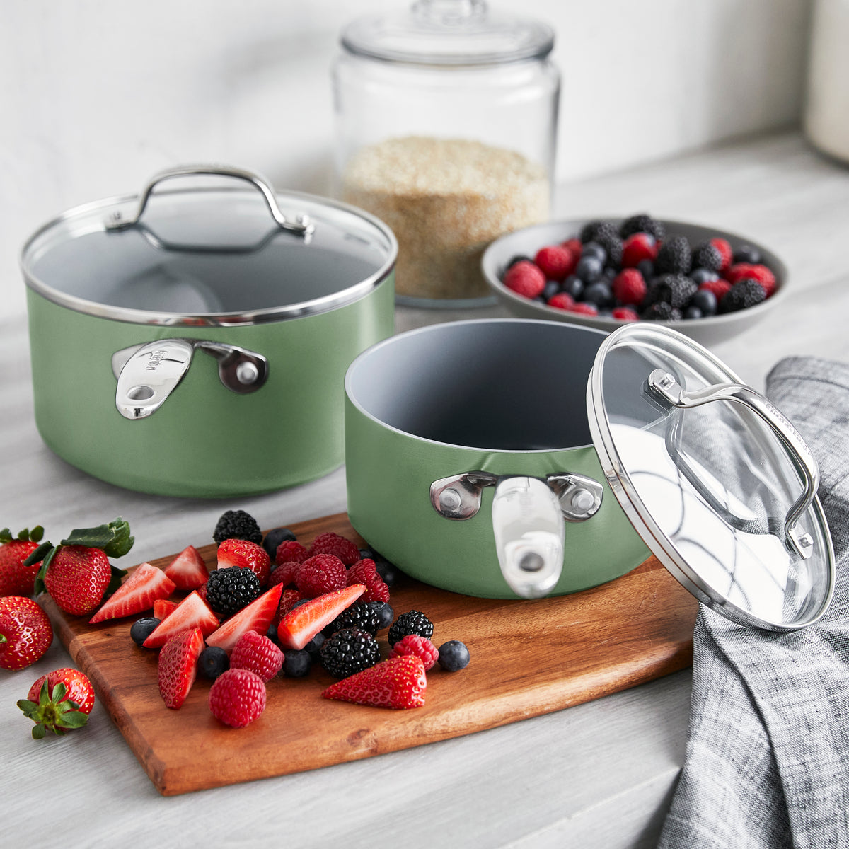 Sage green ceramic nonstick saucepan set with lids on counter, berries and a jar of grains nearby.