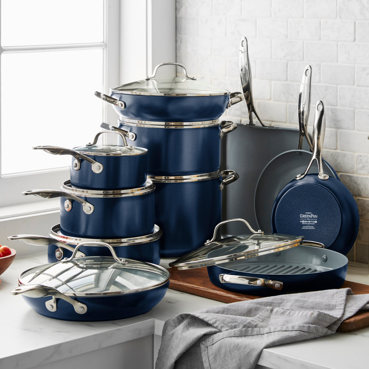 A Valencia Pro Ceramic Nonstick 19-Piece Cookware Set | Twilight is displayed on a white kitchen counter by a window, with some pieces stacked and a gray kitchen towel and cutting board nearby.