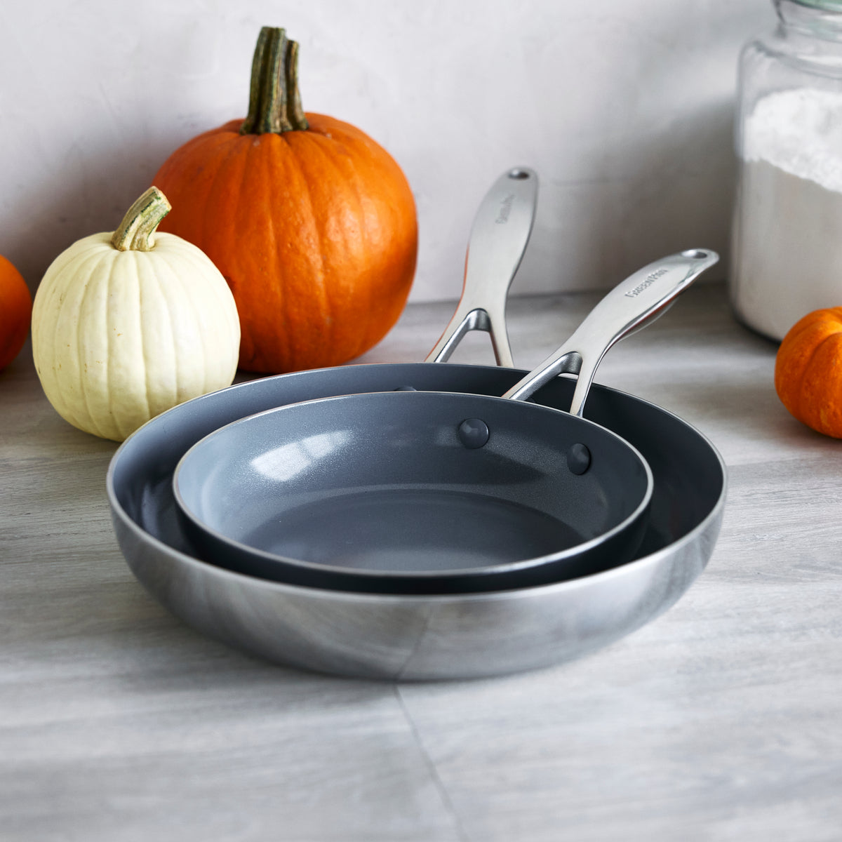 Two stainless steel frypans, 8″ and 10″, with ceramic finish stacked on a counter beside pumpkins and a jar of flour.