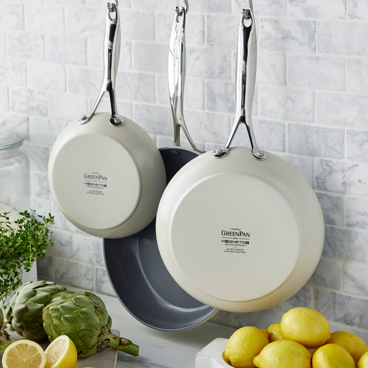 Two green ceramic nonstick skillets hanging on a white tile wall above a counter with lemons, an artichoke, herbs, and a glass jar.