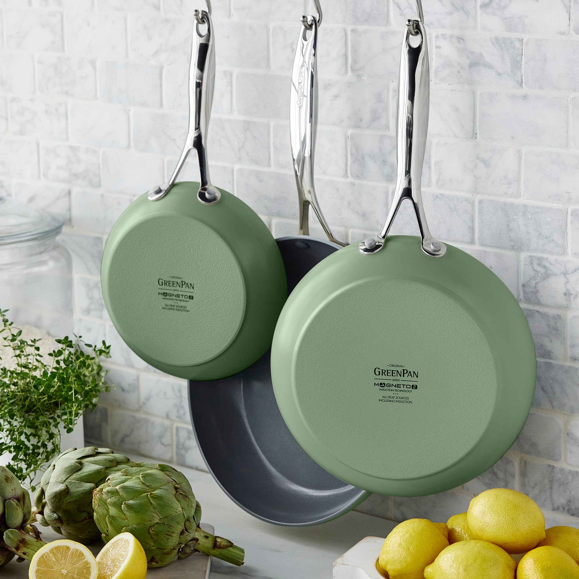 Two Valencia Pro Ceramic Nonstick frypans (Sage), with stainless steel handles, hang on a white tile kitchen wall. Artichokes, lemons, fresh herbs, and a glass jar are in the foreground. Pan sizes: 8", 9.5", and 11".
