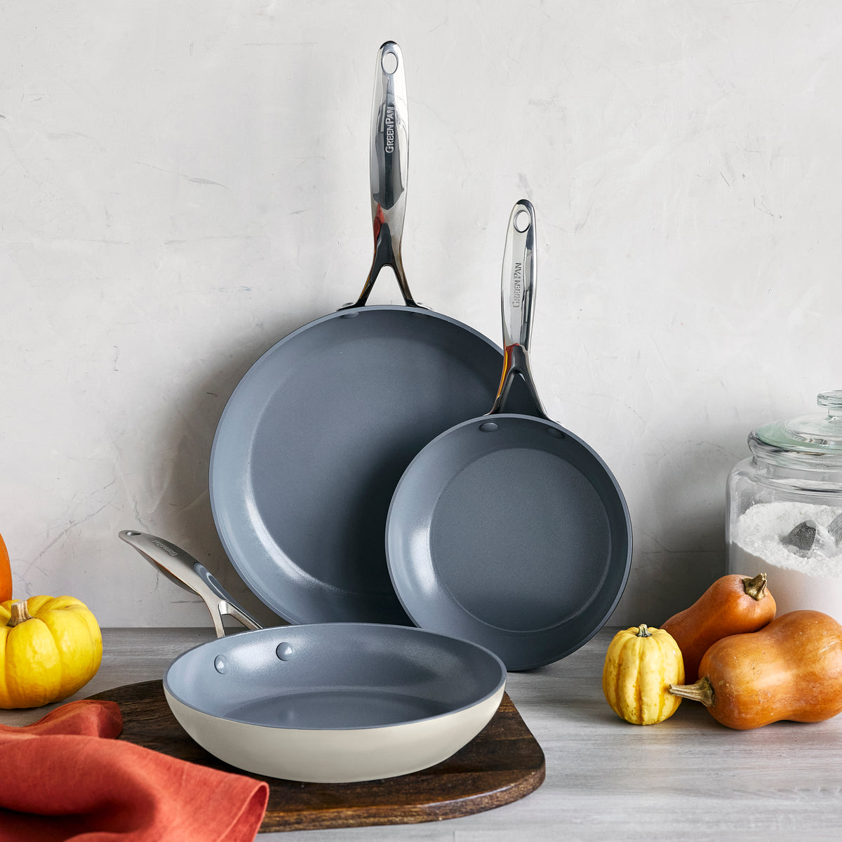 Cream ceramic nonstick frypan set of three on a table with pumpkins, a flour jar, and an orange napkin.