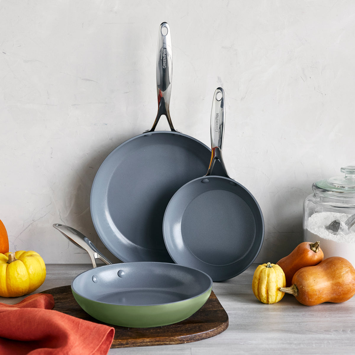 Set of three sage green ceramic nonstick frypans on a counter with pumpkins, squash, a sugar jar, and an orange cloth.