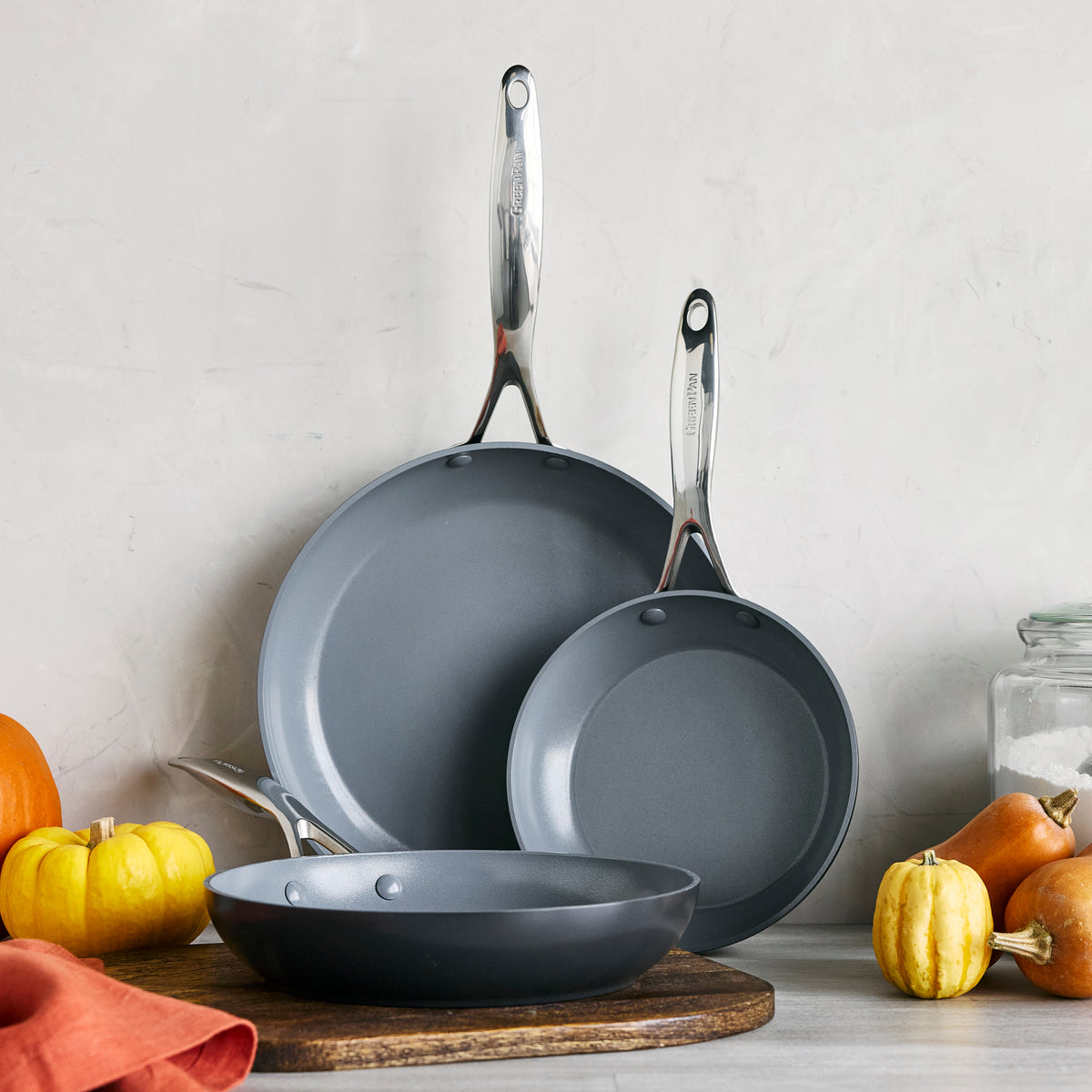 Three gray ceramic nonstick frypans stacked upright on a counter with pumpkins and gourds.