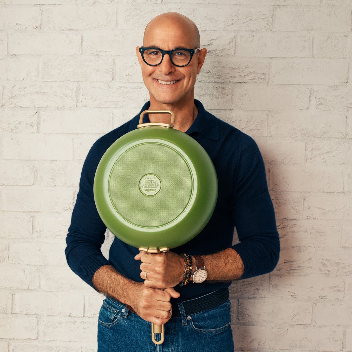 Stanley Tucci holding a green pan against a light brick wall