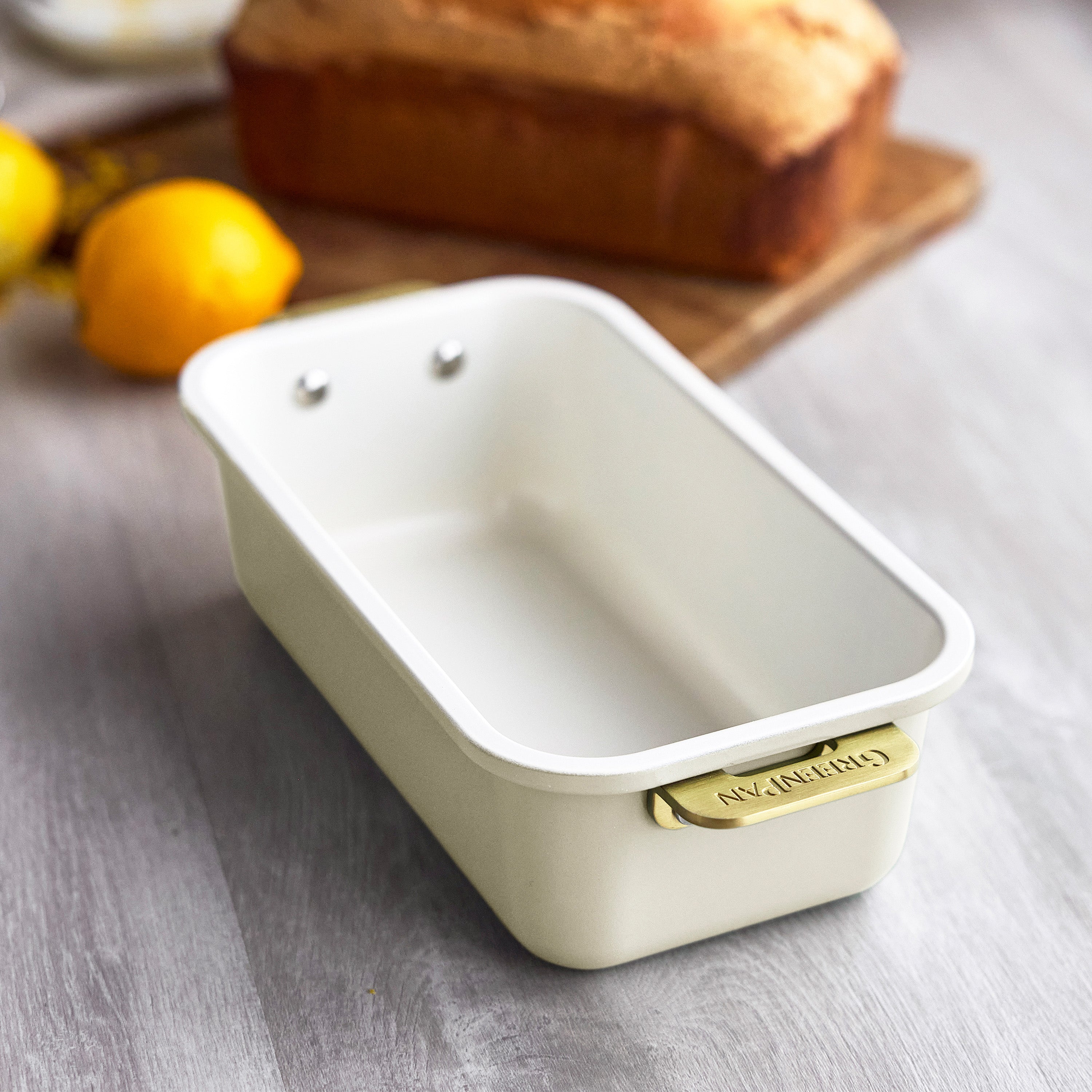 Cream loaf pan with gold handle on a gray surface, with lemons and bread in the background.