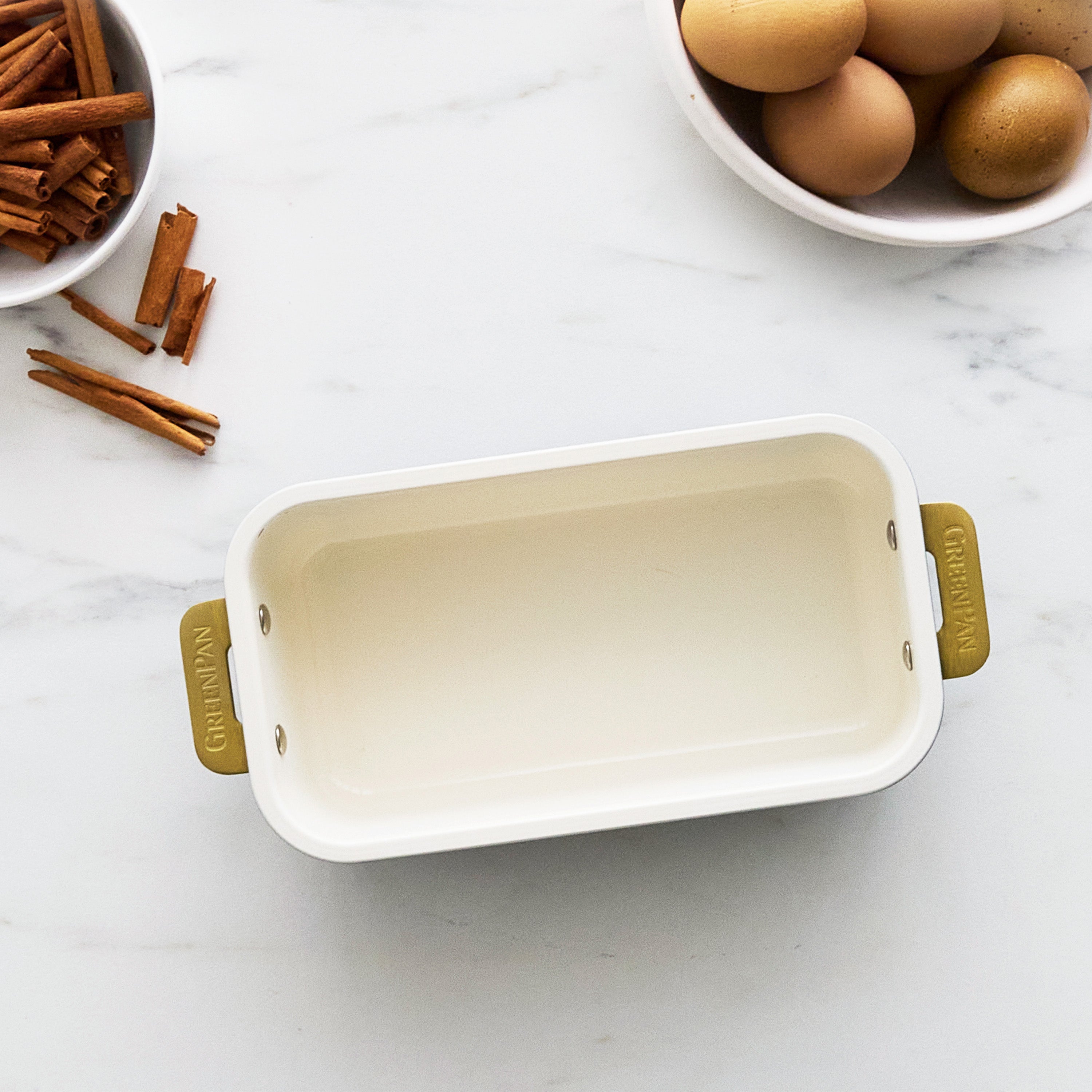Cream loaf pan with gold handles on a marble surface with cinnamon sticks and eggs.