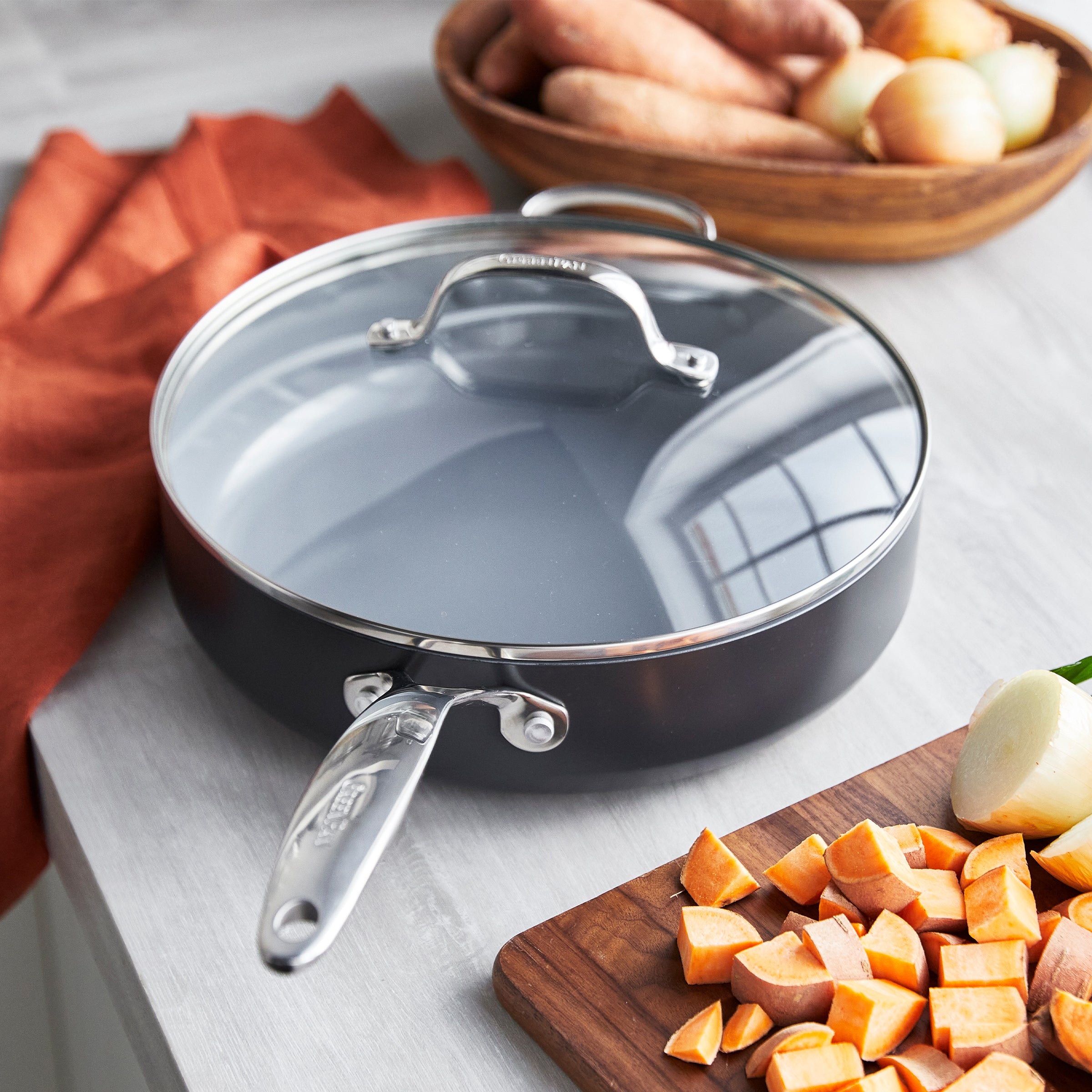 Ceramic nonstick sauté pan with lid next to sweet potatoes, red onions, and assorted vegetables on a kitchen surface.