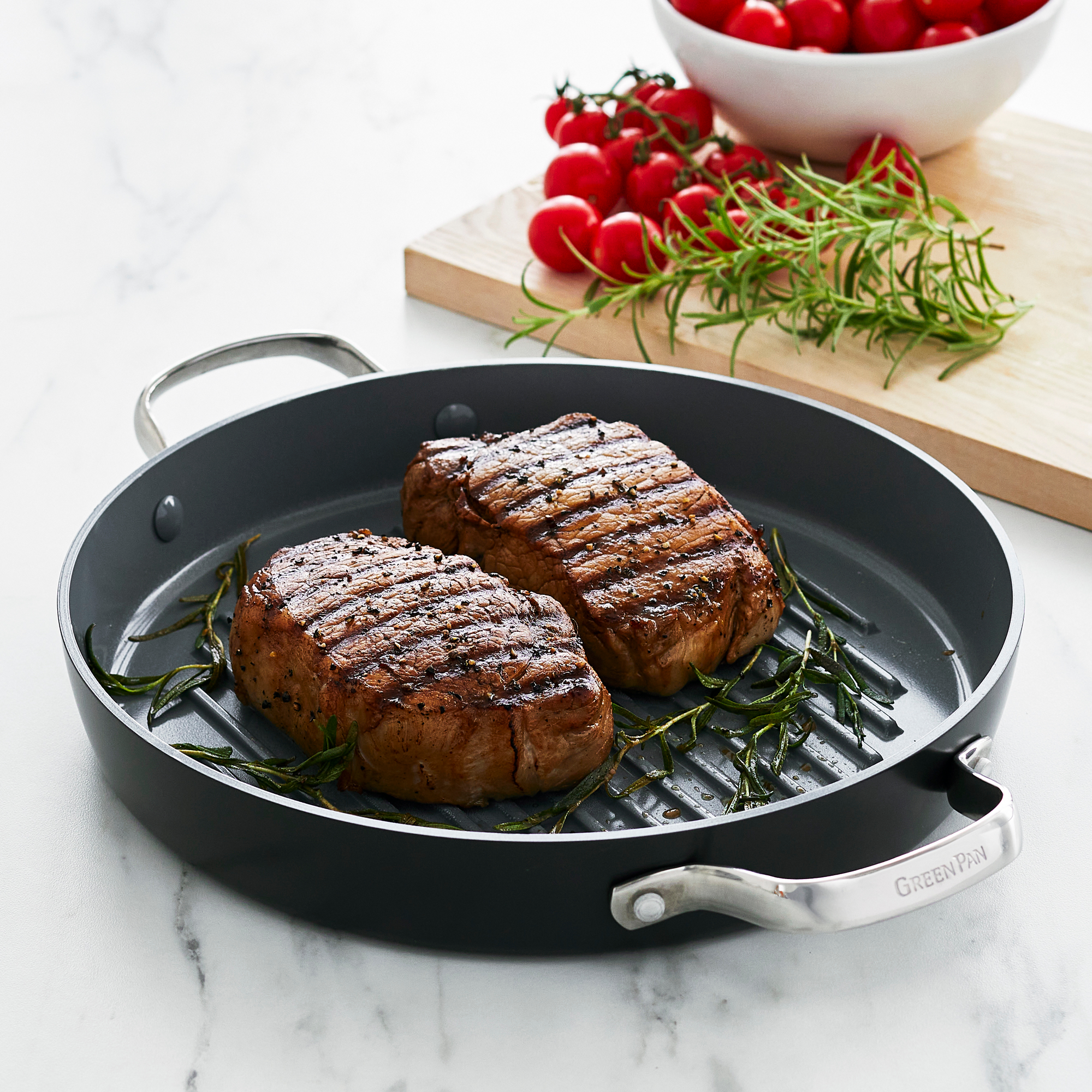 Two grilled steaks with rosemary in a gray ceramic grill pan, cherry tomatoes and herbs on the side.