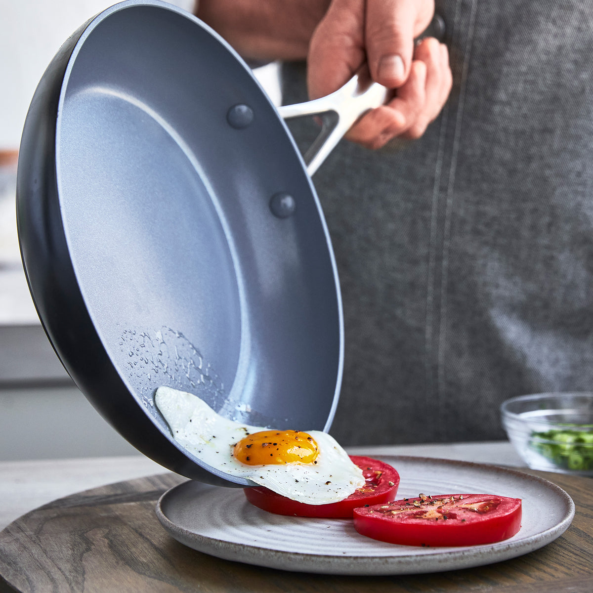 A hand slides a fried egg from a gray Valencia Pro frypan onto a plate with sliced tomatoes.