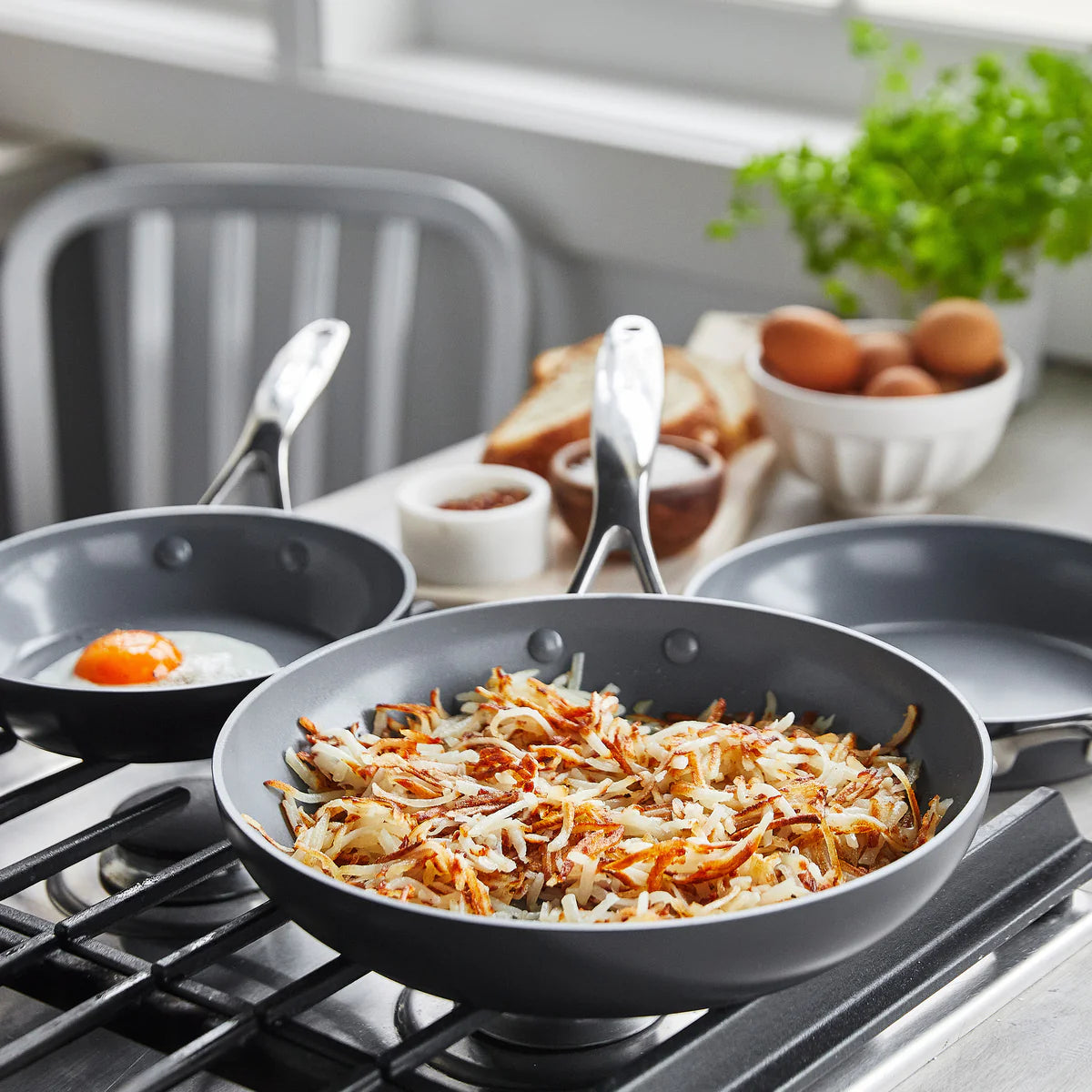 Set of three metal pans on a stovetop, one with hash browns, one with a fried egg, and one empty; eggs and bread in the background.