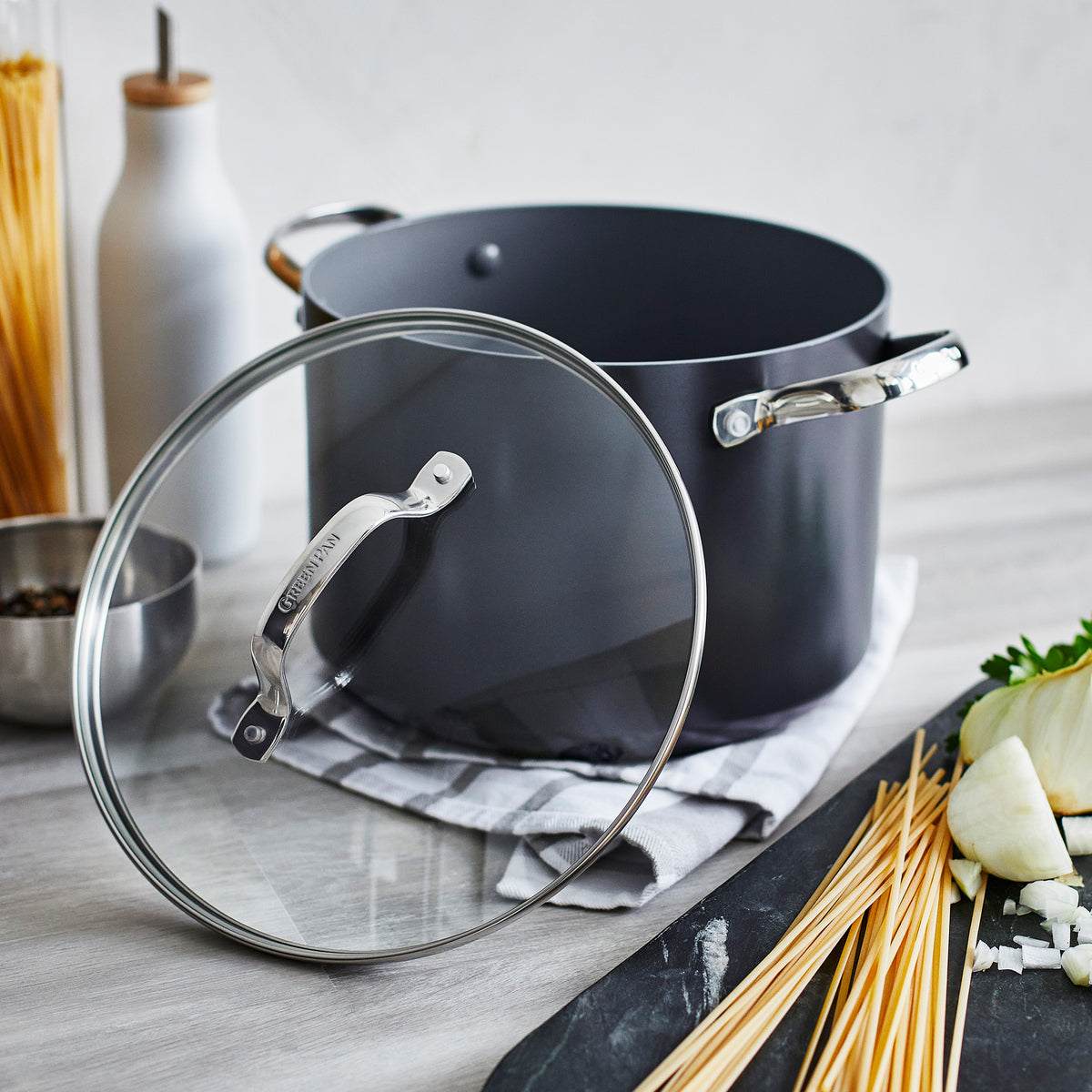 Gray pot with glass lid from a cookware set, surrounded by uncooked spaghetti, onion, parsley, and a glass bottle.