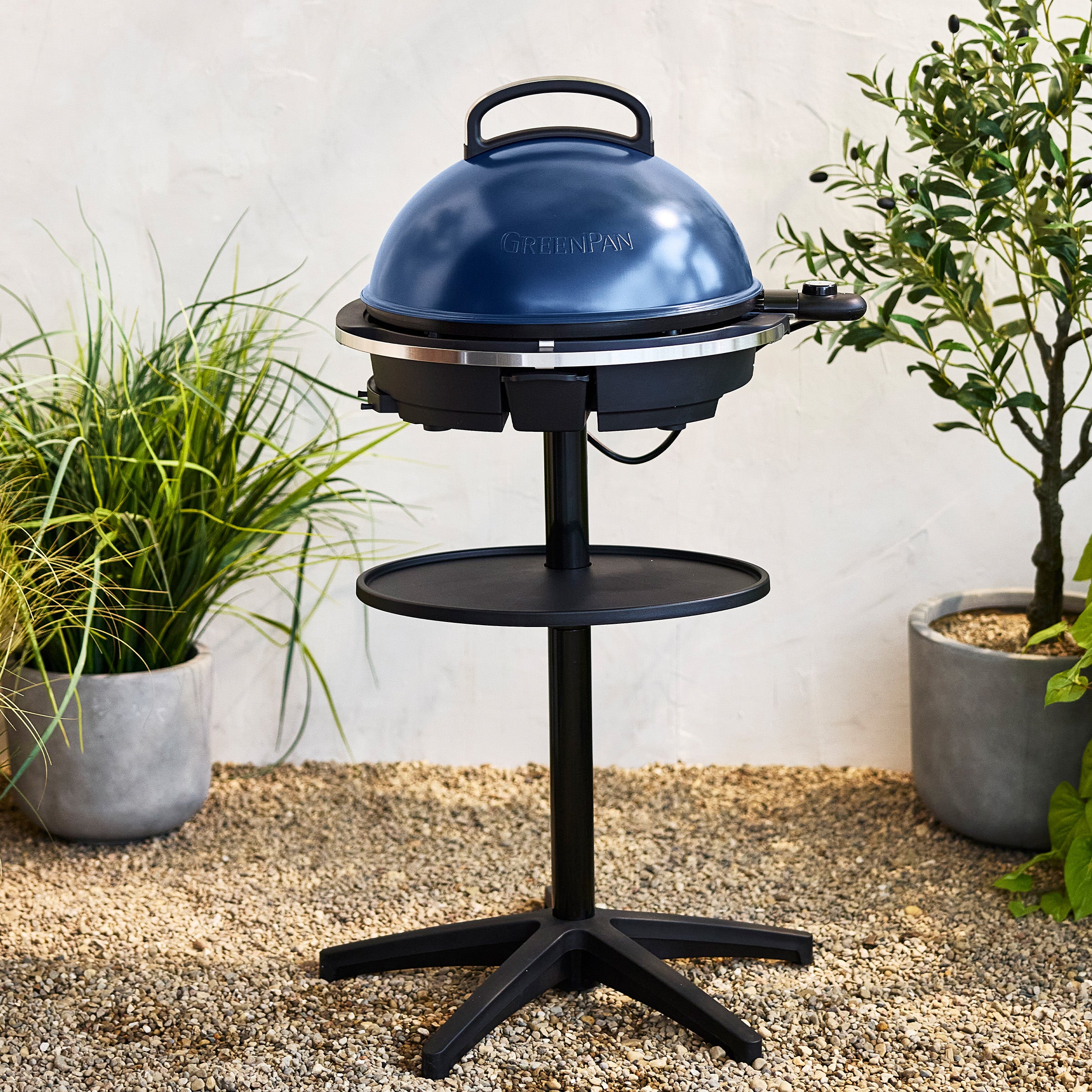Dark blue portable grill on a stand with plants in the background