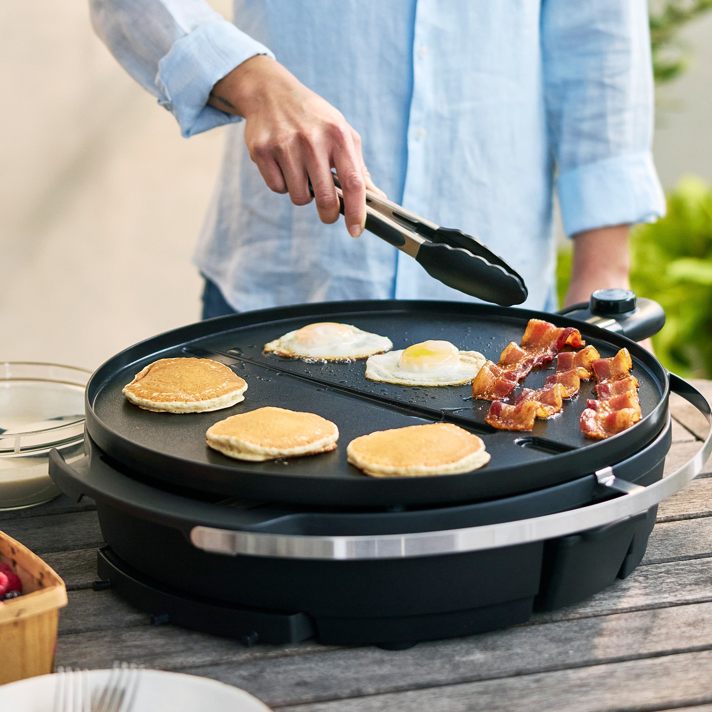 Person cooking pancakes, eggs and bacon on a portable griddle outdoors.