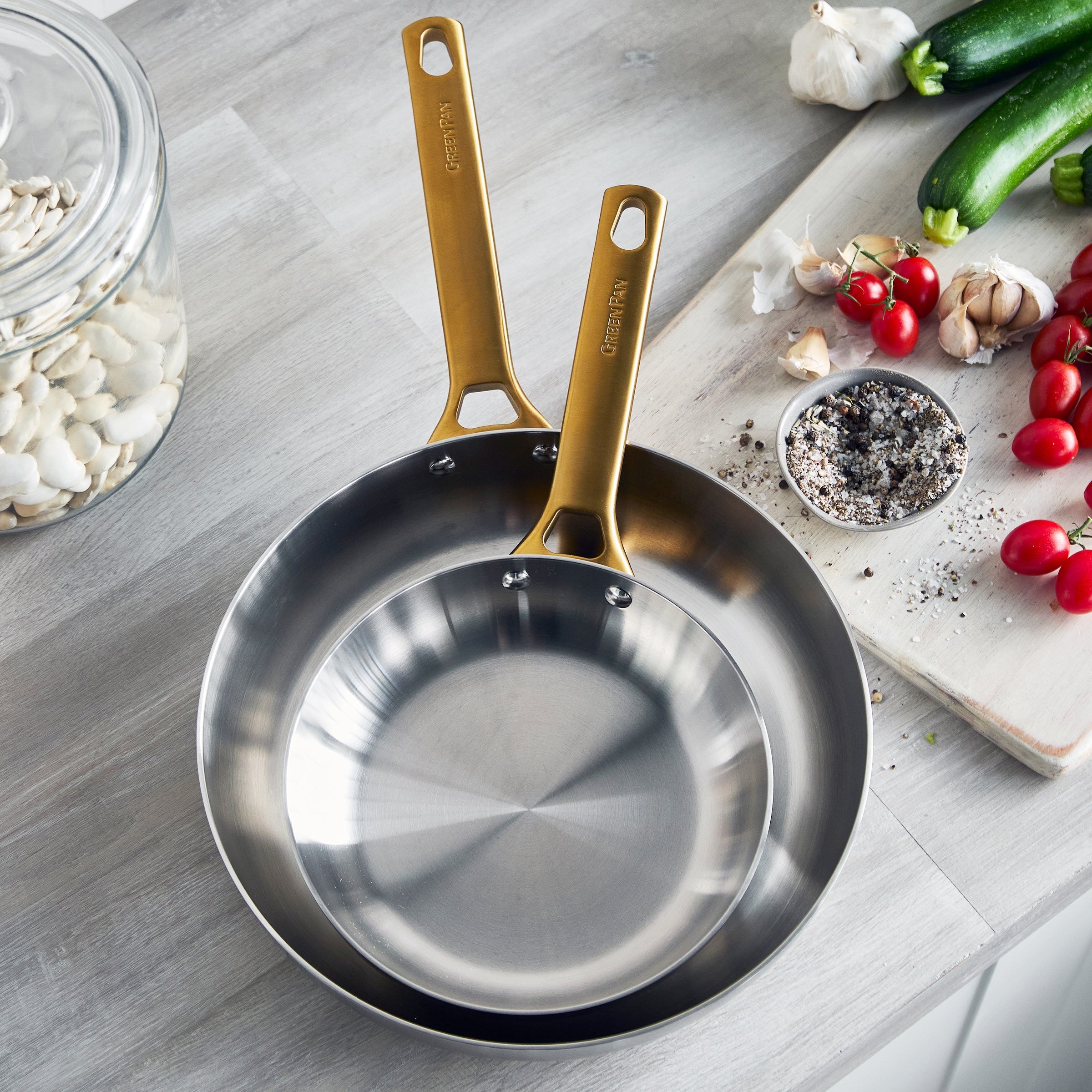 Stainless steel frying pans with gold handles on a kitchen counter with vegetables and spices.