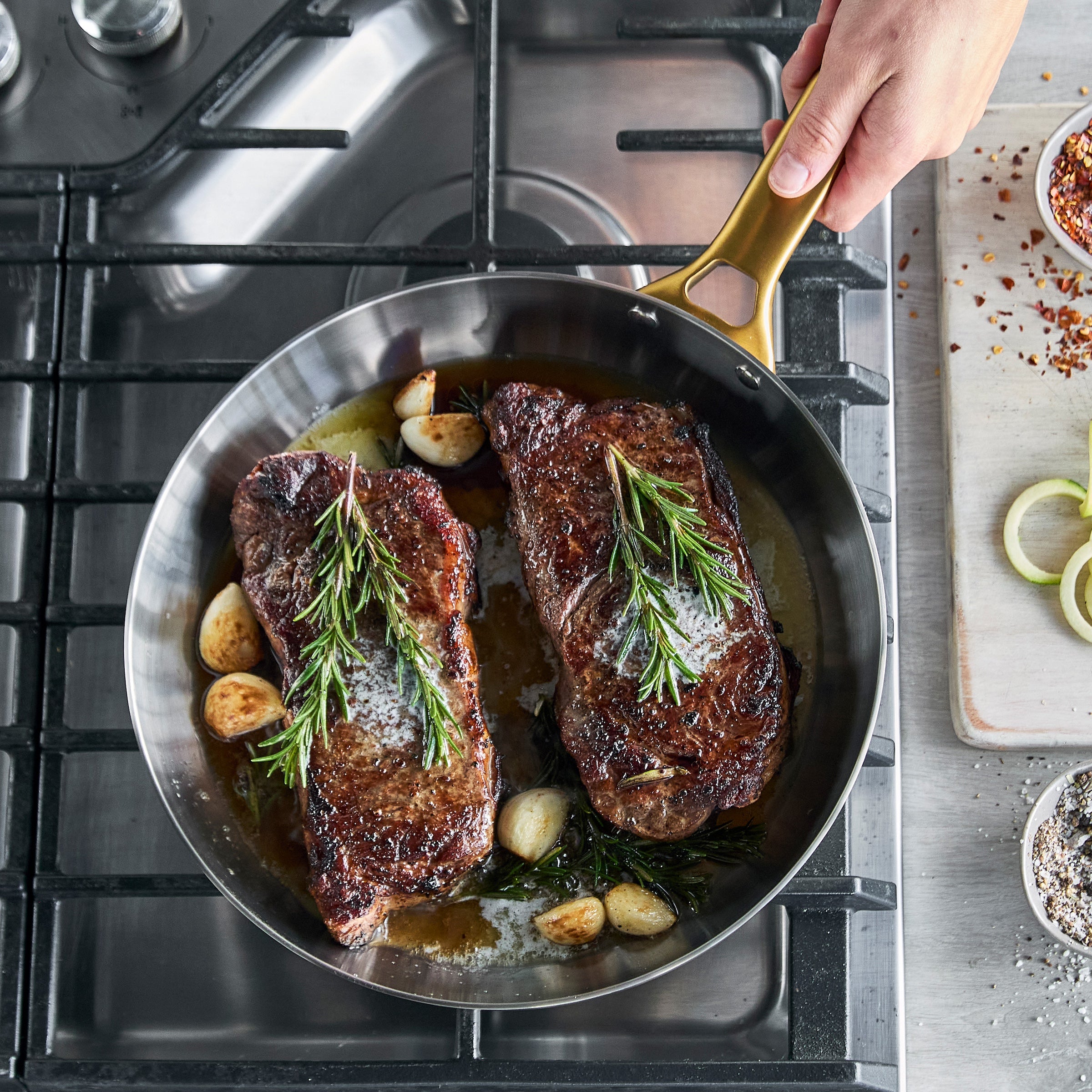 Two steaks with rosemary and garlic in a frying pan on a stovetop.