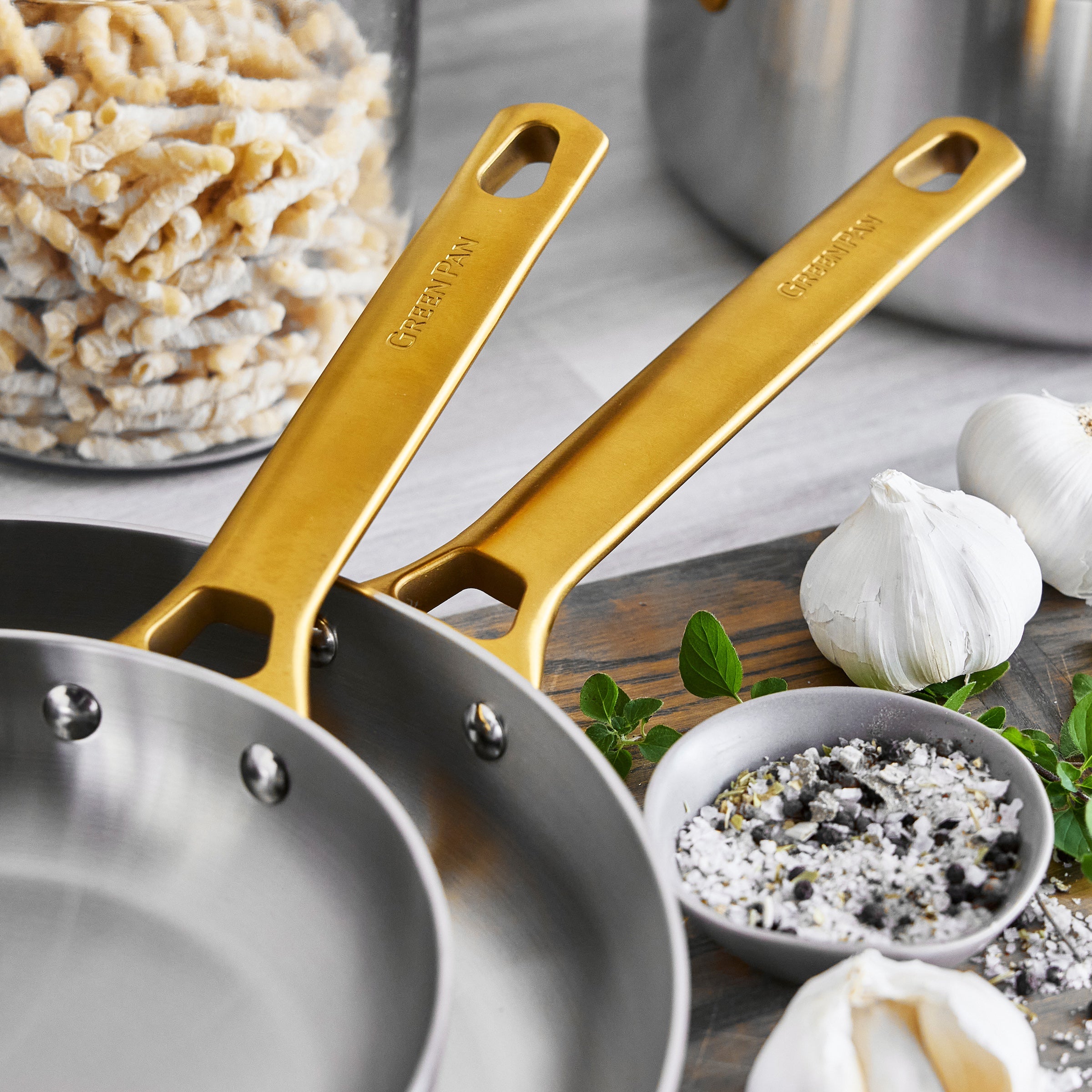 Two stainless steel frypans with gold handles on a kitchen counter with ingredients.