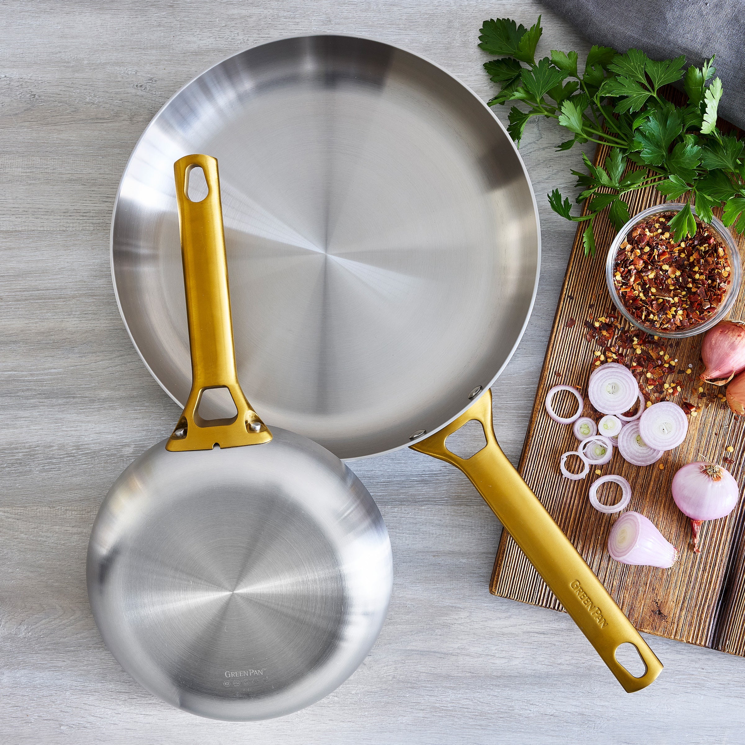 Two stainless steel frying pans with gold handles on a gray surface with ingredients.