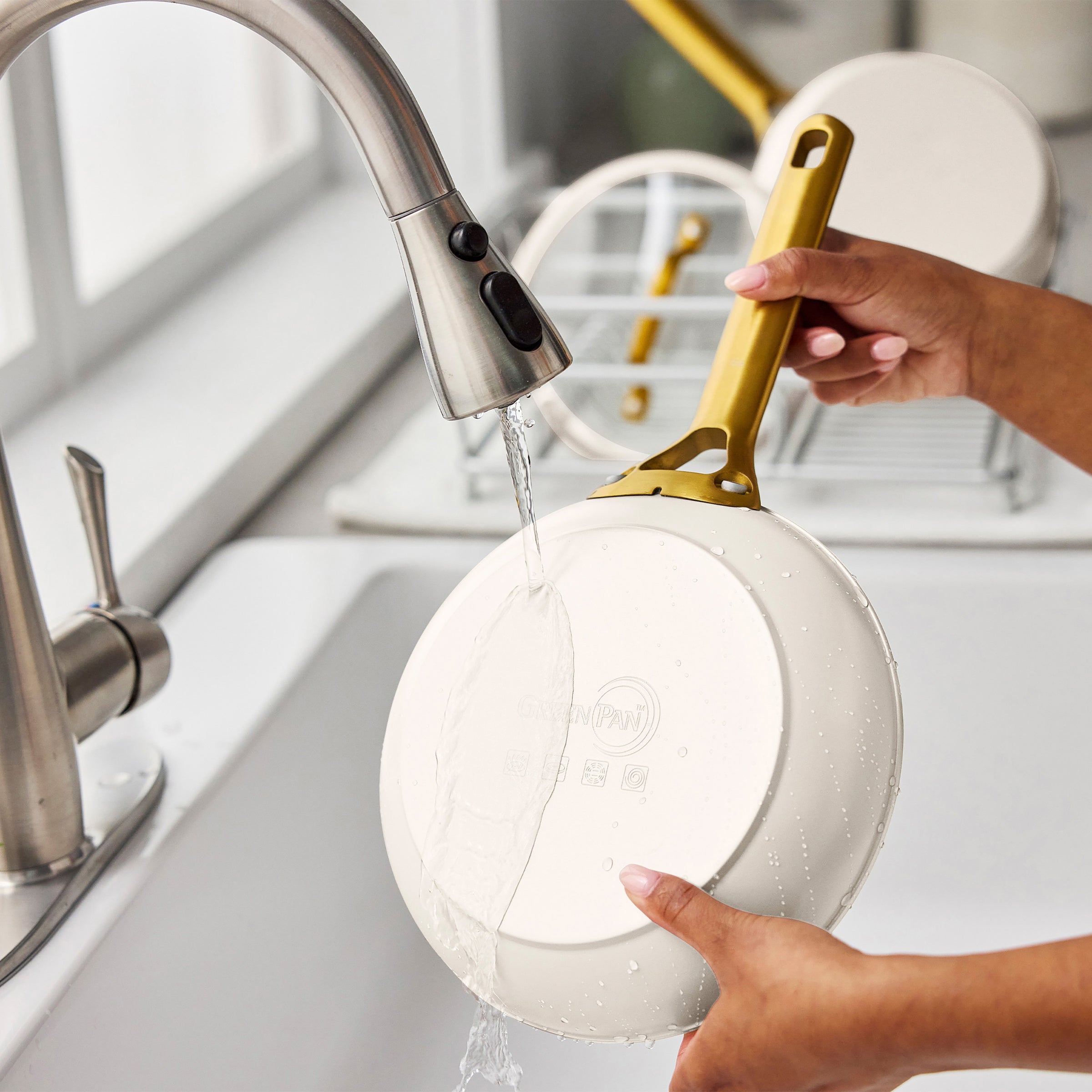 Person washing a cream frying pan with a gold handle under running water in a kitchen sink.