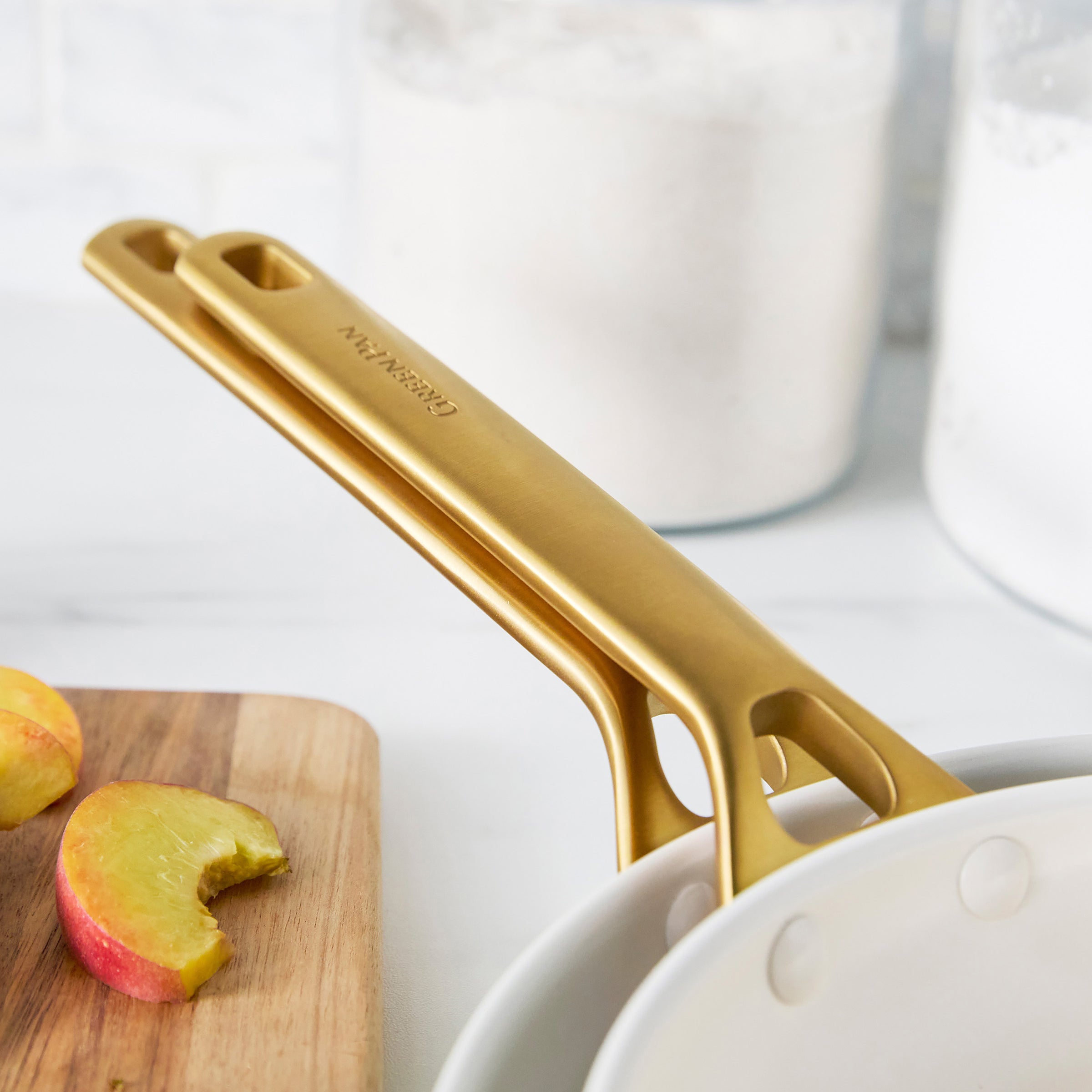 Gold frypan handles on a white surface with apples and a cutting board.
