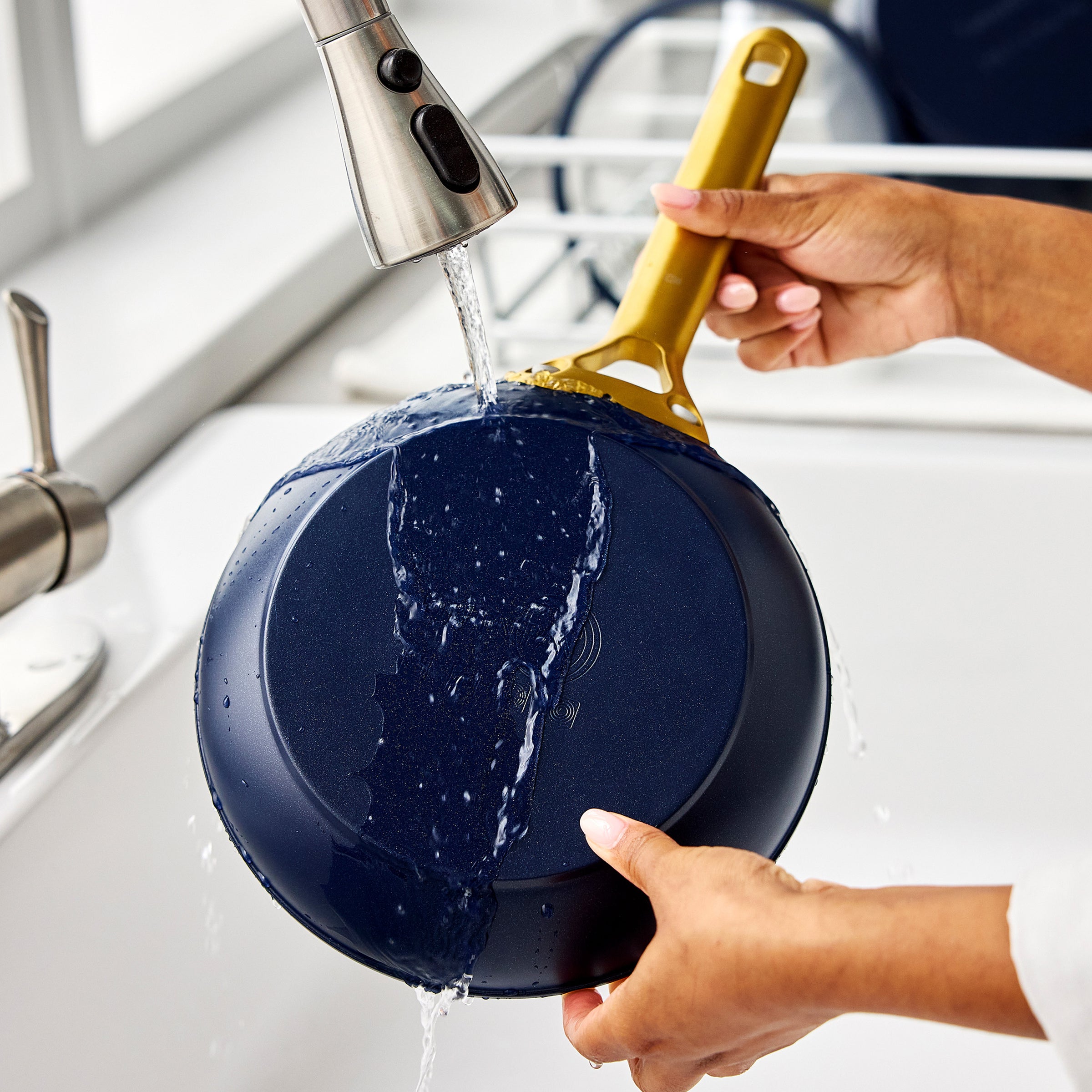 Person washing a dark blue frying pan under running water in a kitchen sink