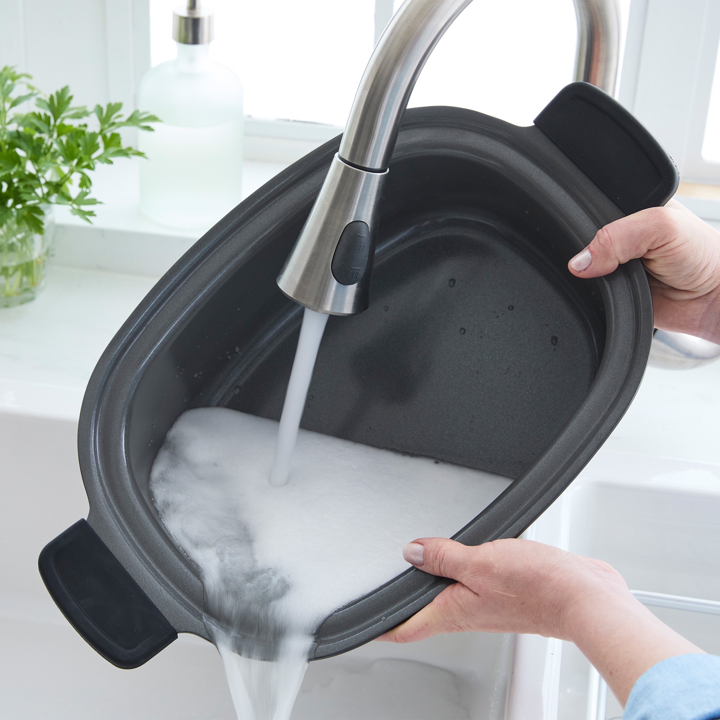 Person washing a removable slow cooker pot in a sink with running water.