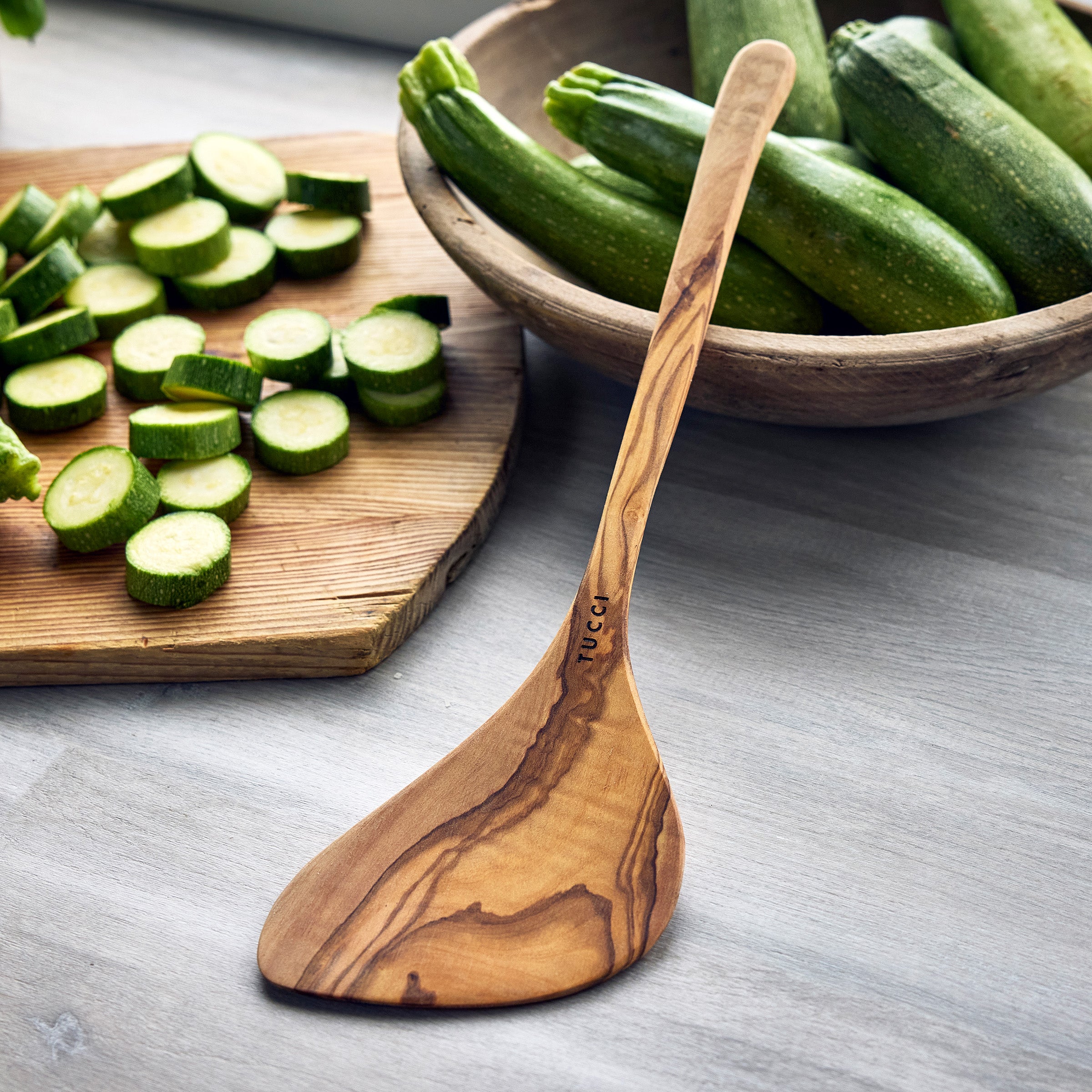 Wooden spoon on a table with sliced zucchinis and a bowl of whole zucchinis in the background