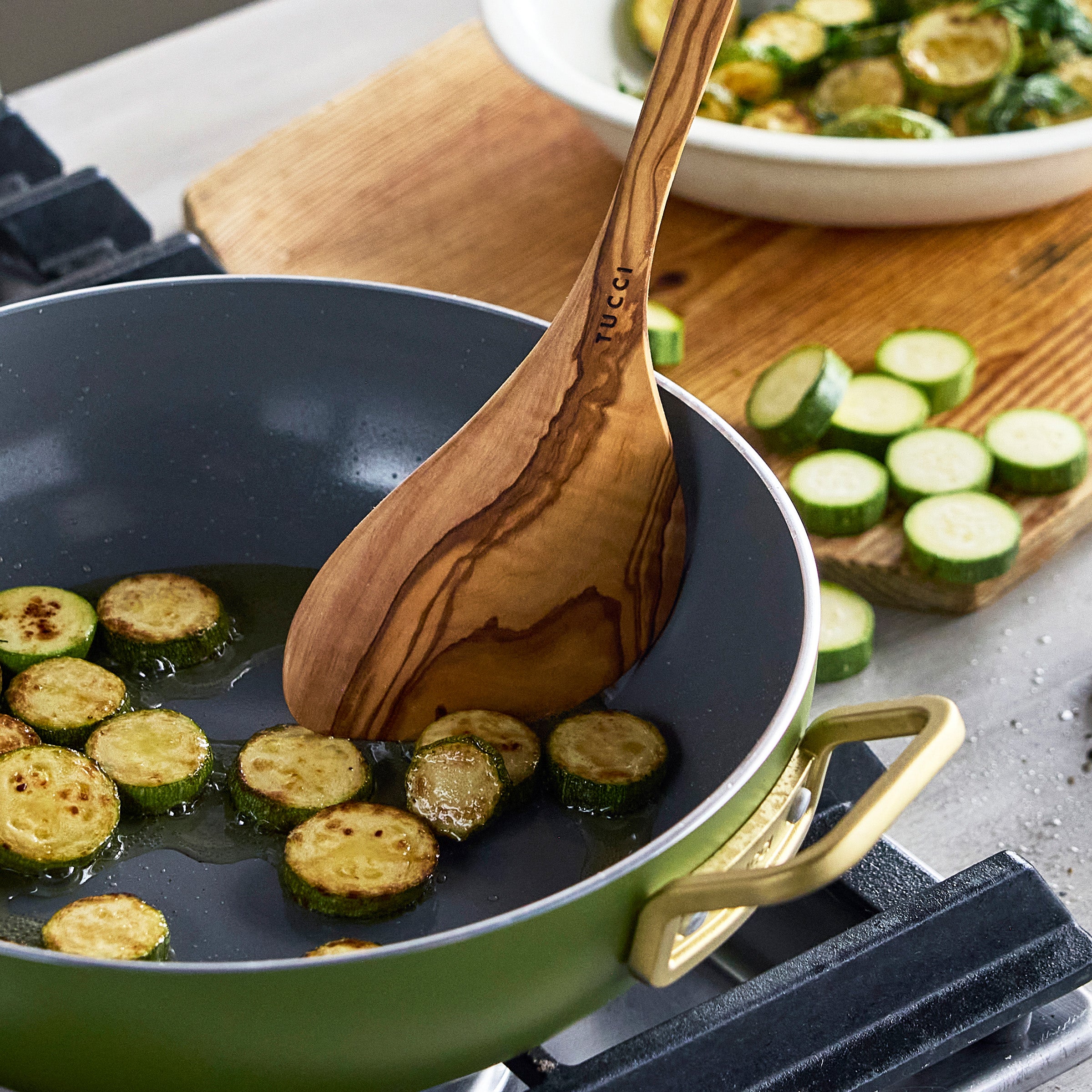 Zucchini being cooked in a green frying pan with a wooden spoon