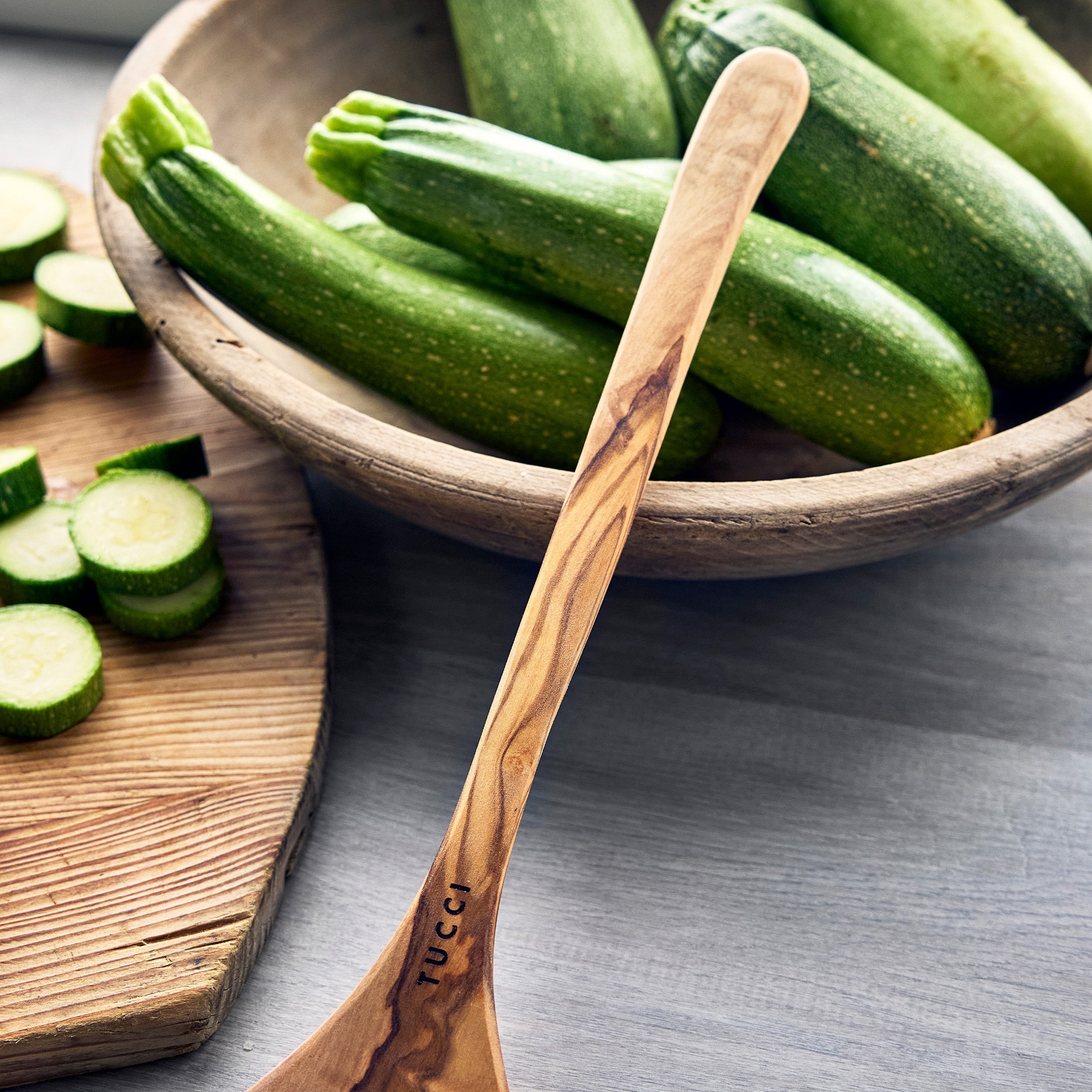Wooden spoon with 'Tucci' branding in front of a bowl of green zucchinis on a gray surface