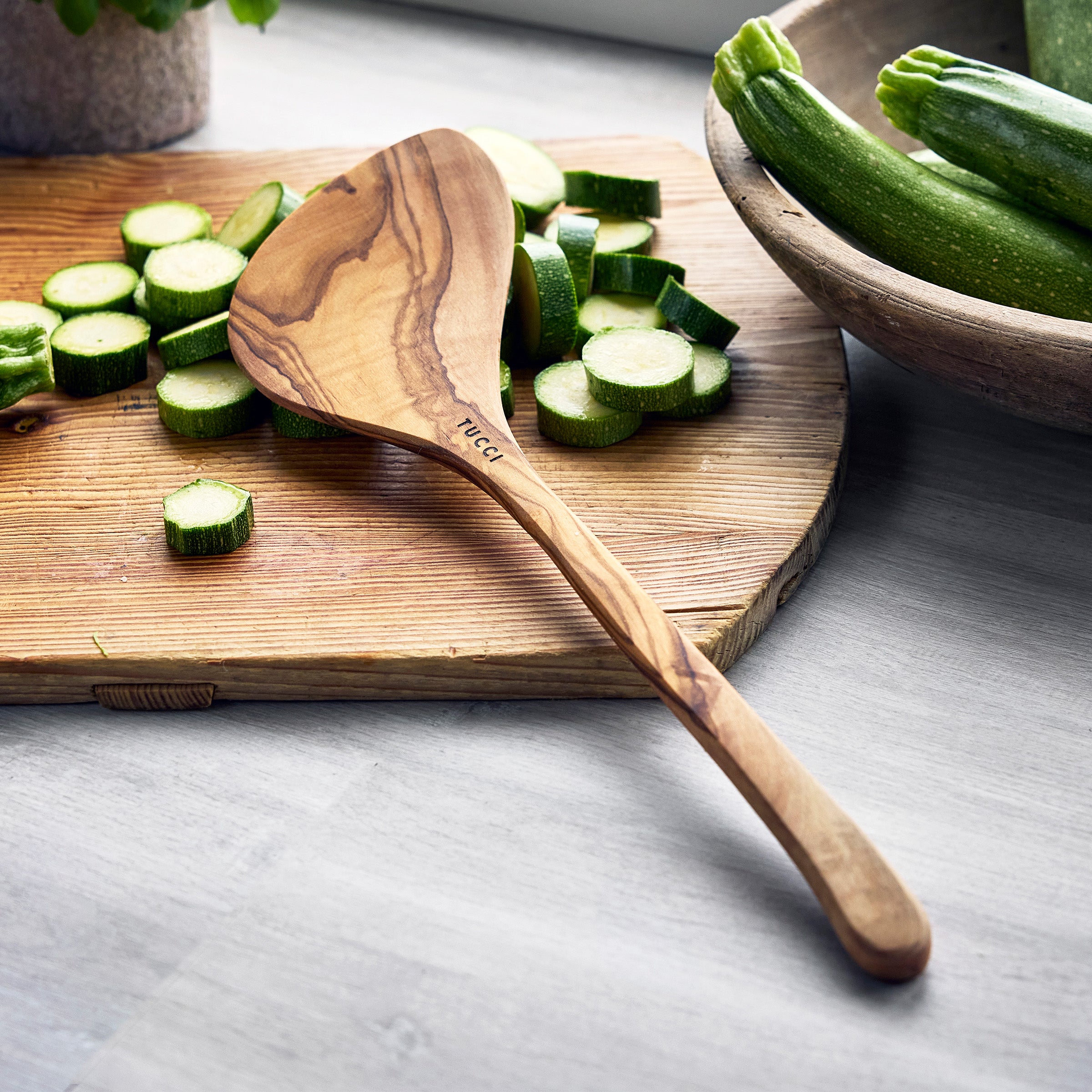 Wooden spoon on a cutting board with sliced zucchinis and a bowl of zucchinis