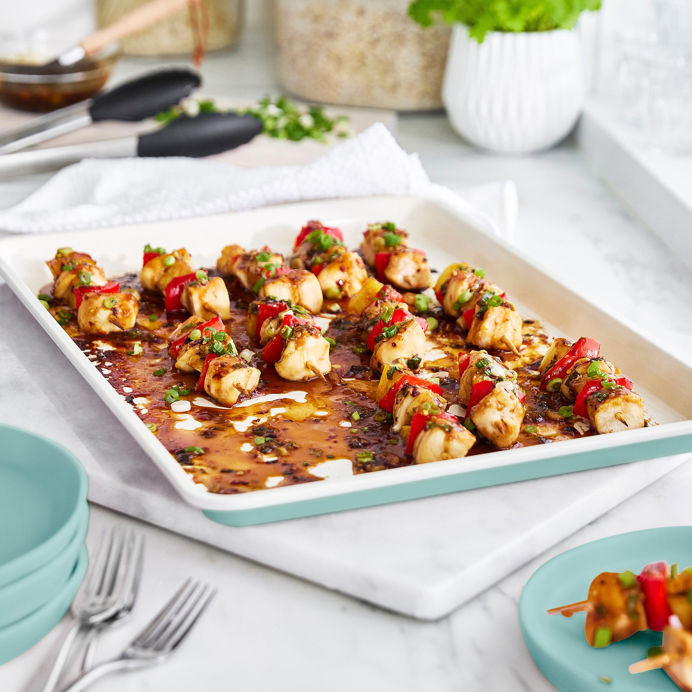 Sheet pan of skewered chicken and vegetables on a white surface with utensils and plates.