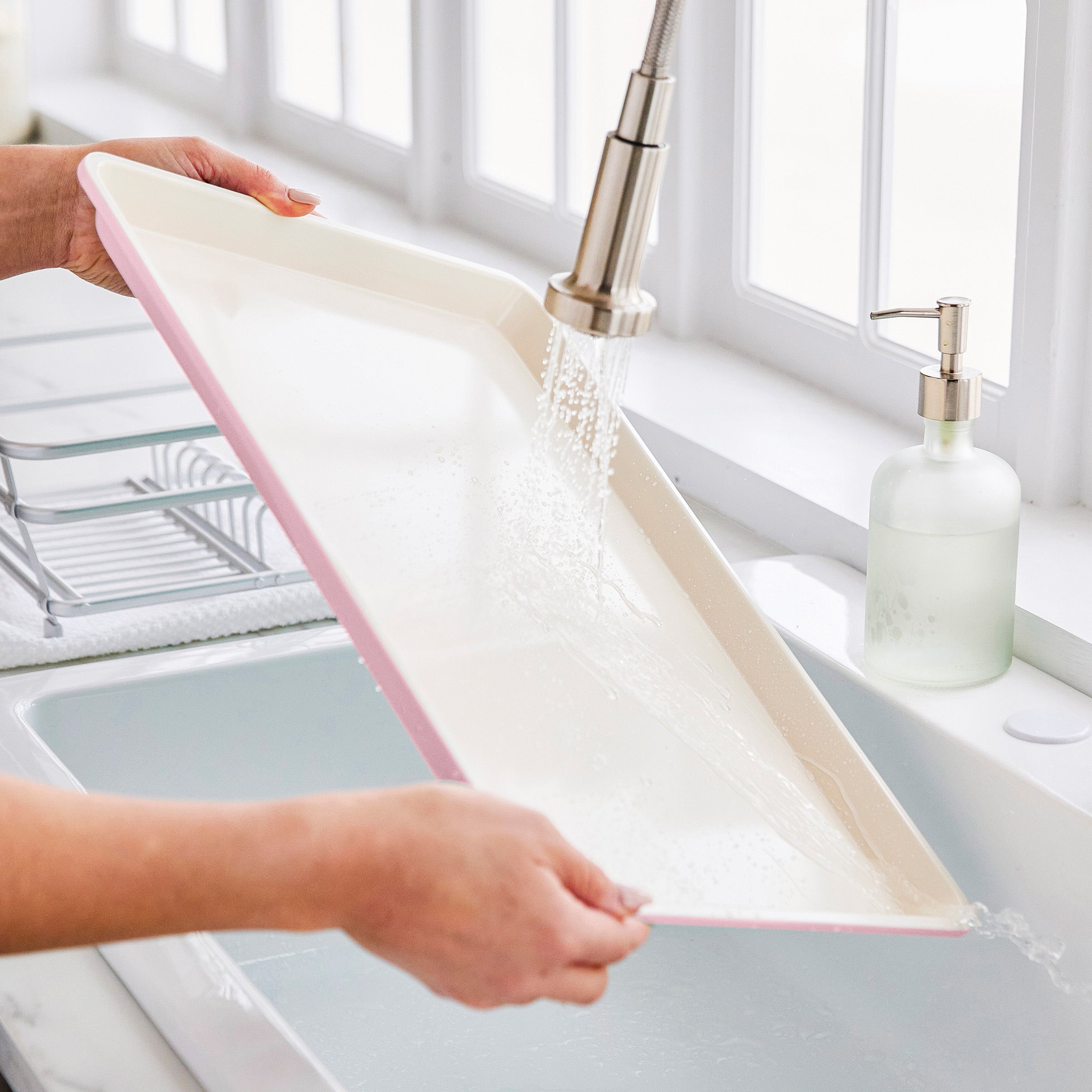 Person washing a pink sheet pan under running water in a kitchen sink.