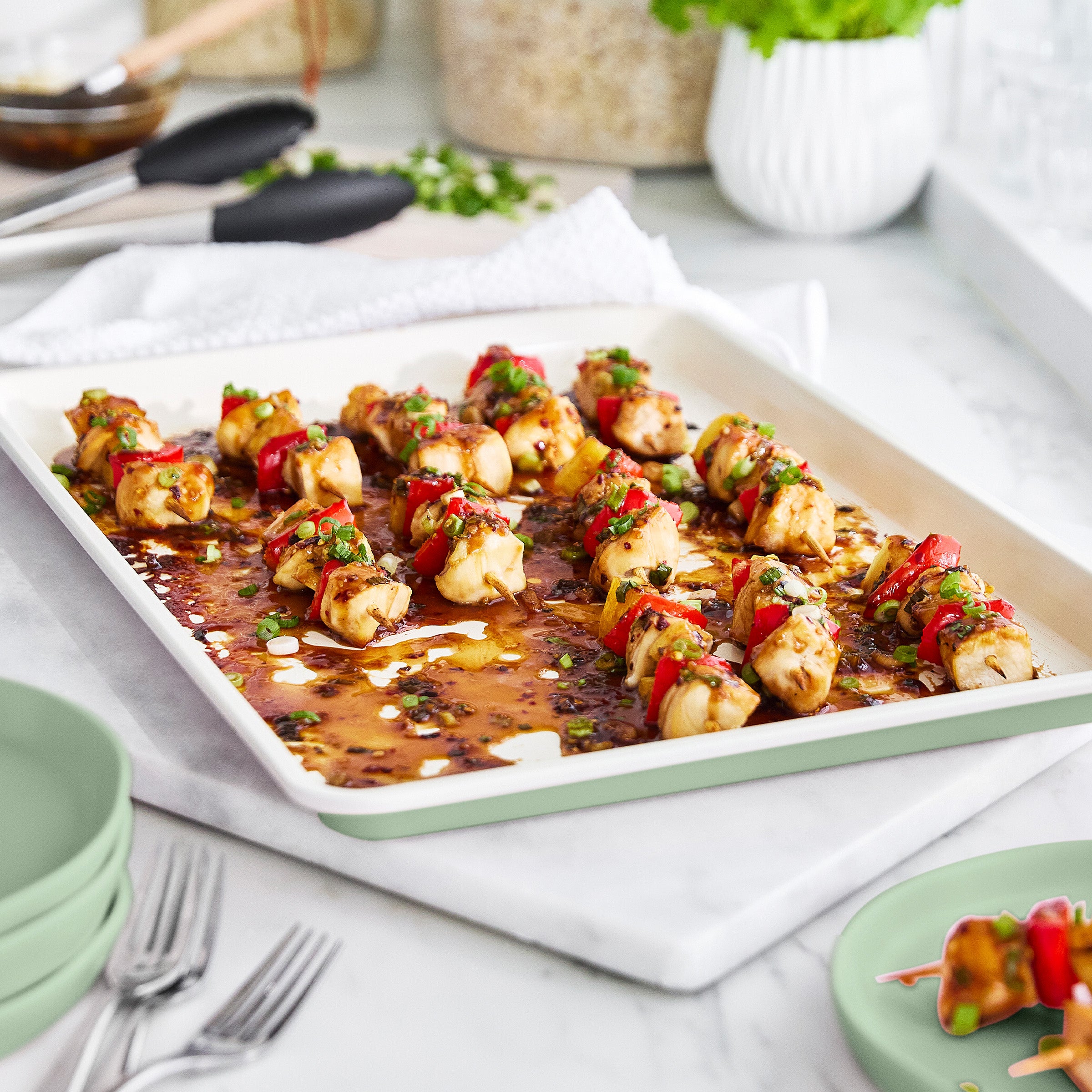 Sheet pan of skewered chicken and vegetables on a white surface with utensils and plates.