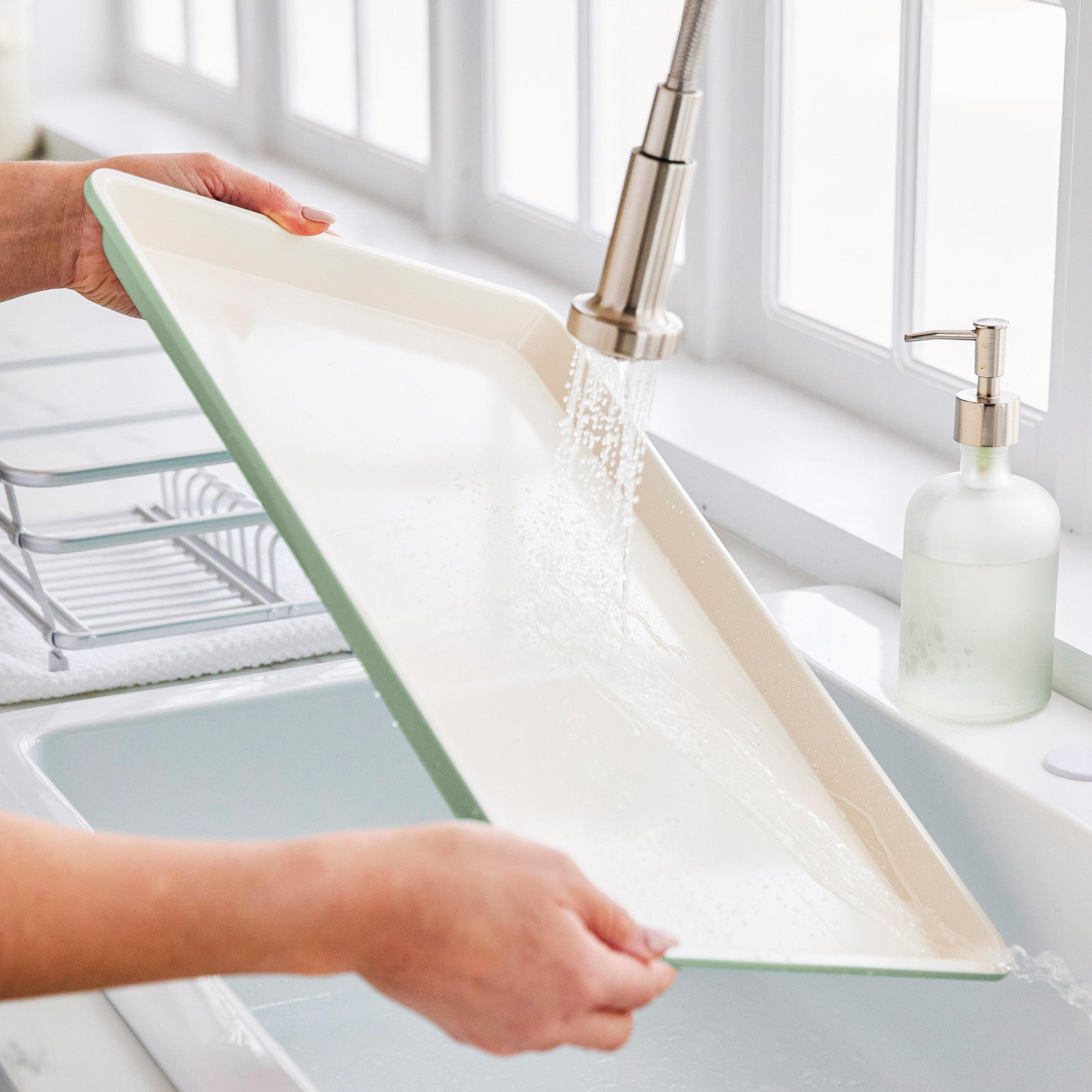 Person washing a green sheet pan under running water in a kitchen sink.
