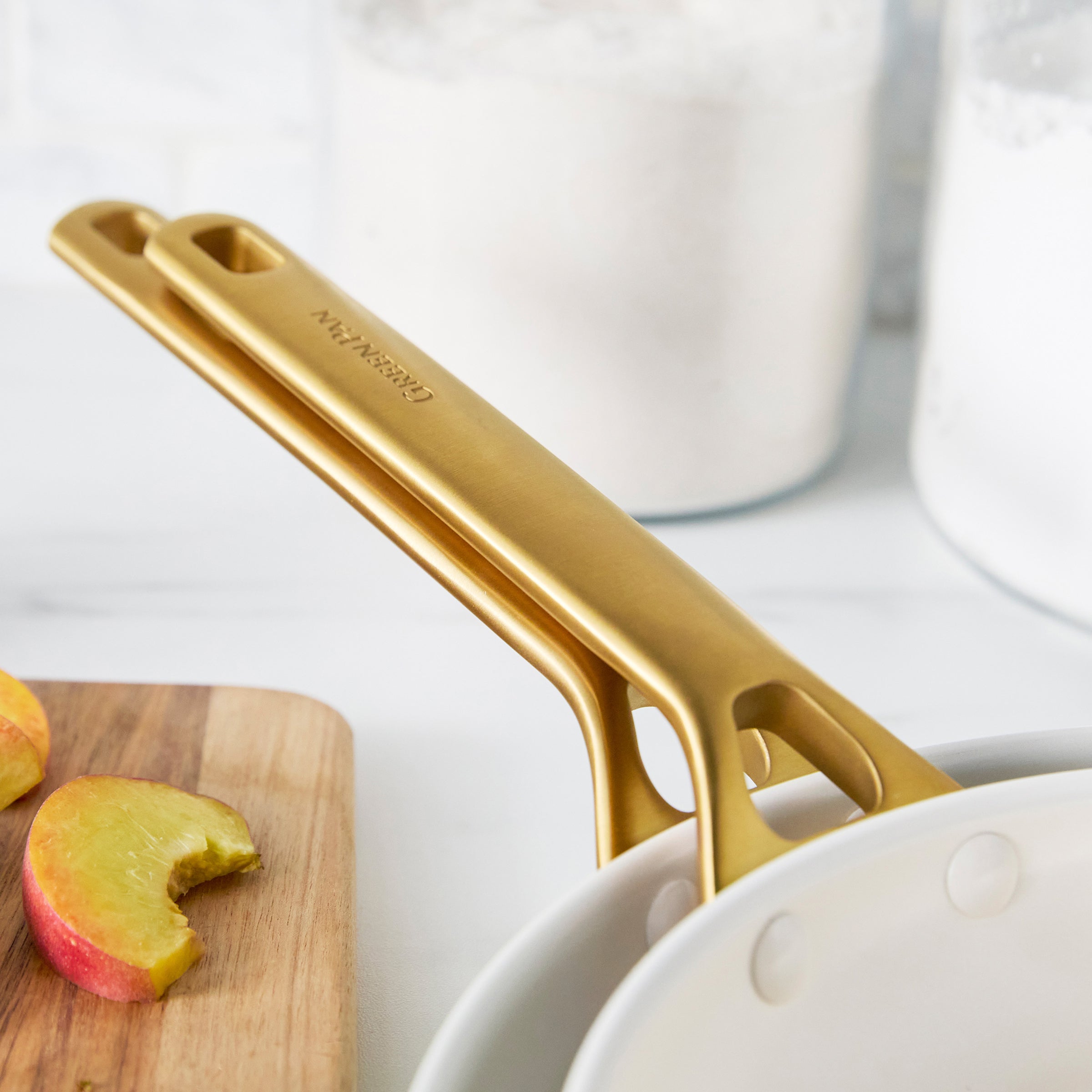 Light blue frypans with gold handles on a white surface with apples and a cutting board in the background