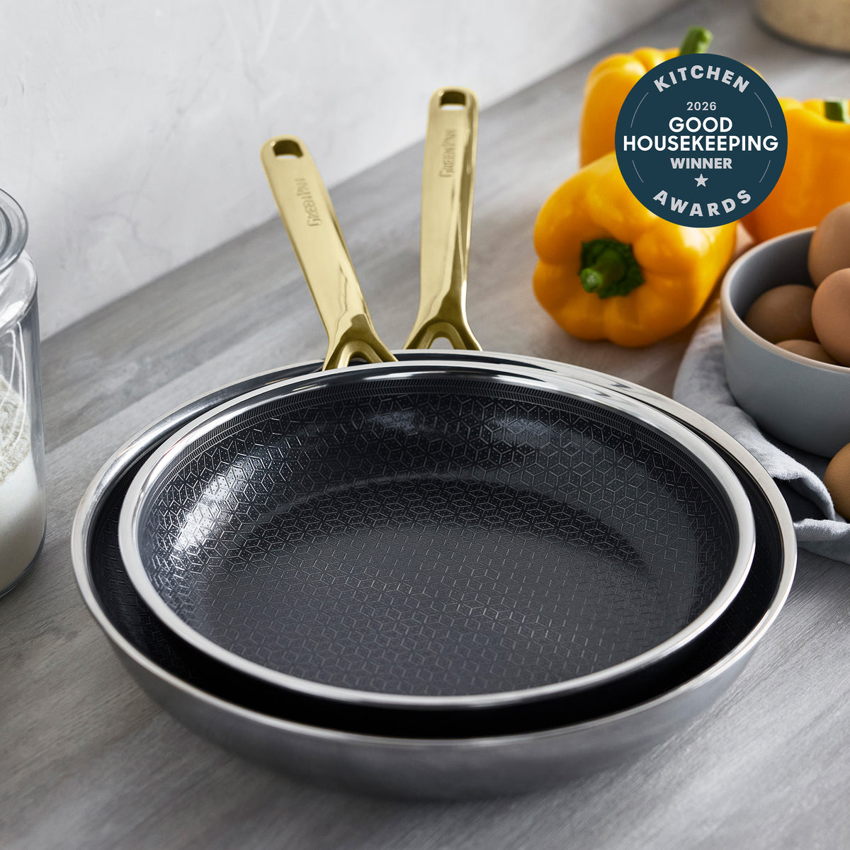 Two gold-handled ceramic frypans, 10″ and 12″, on a surface with eggs, peppers, and an award badge nearby.