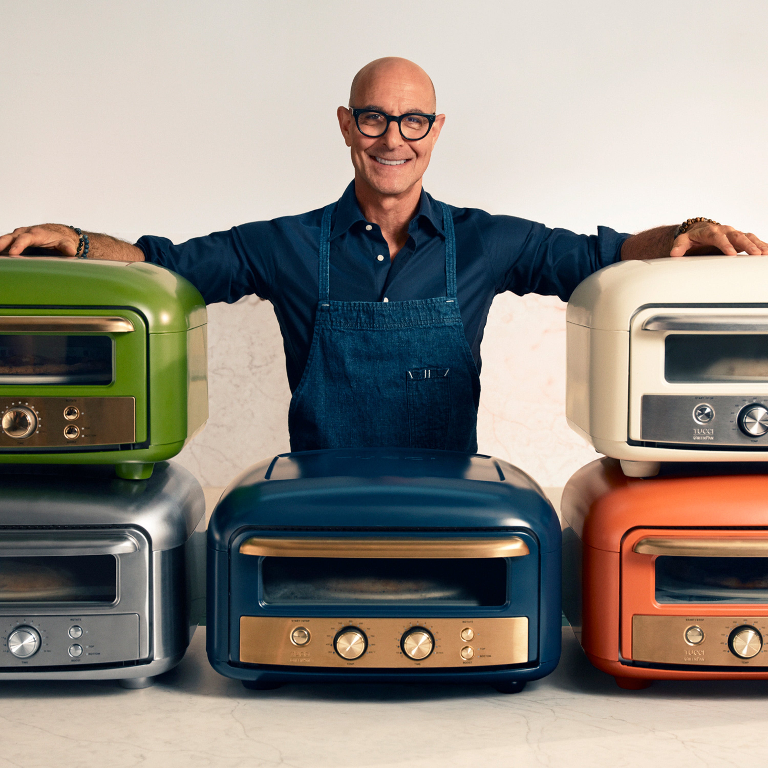 Stanley Tucci in an apron standing behind pizza ovens on a white background