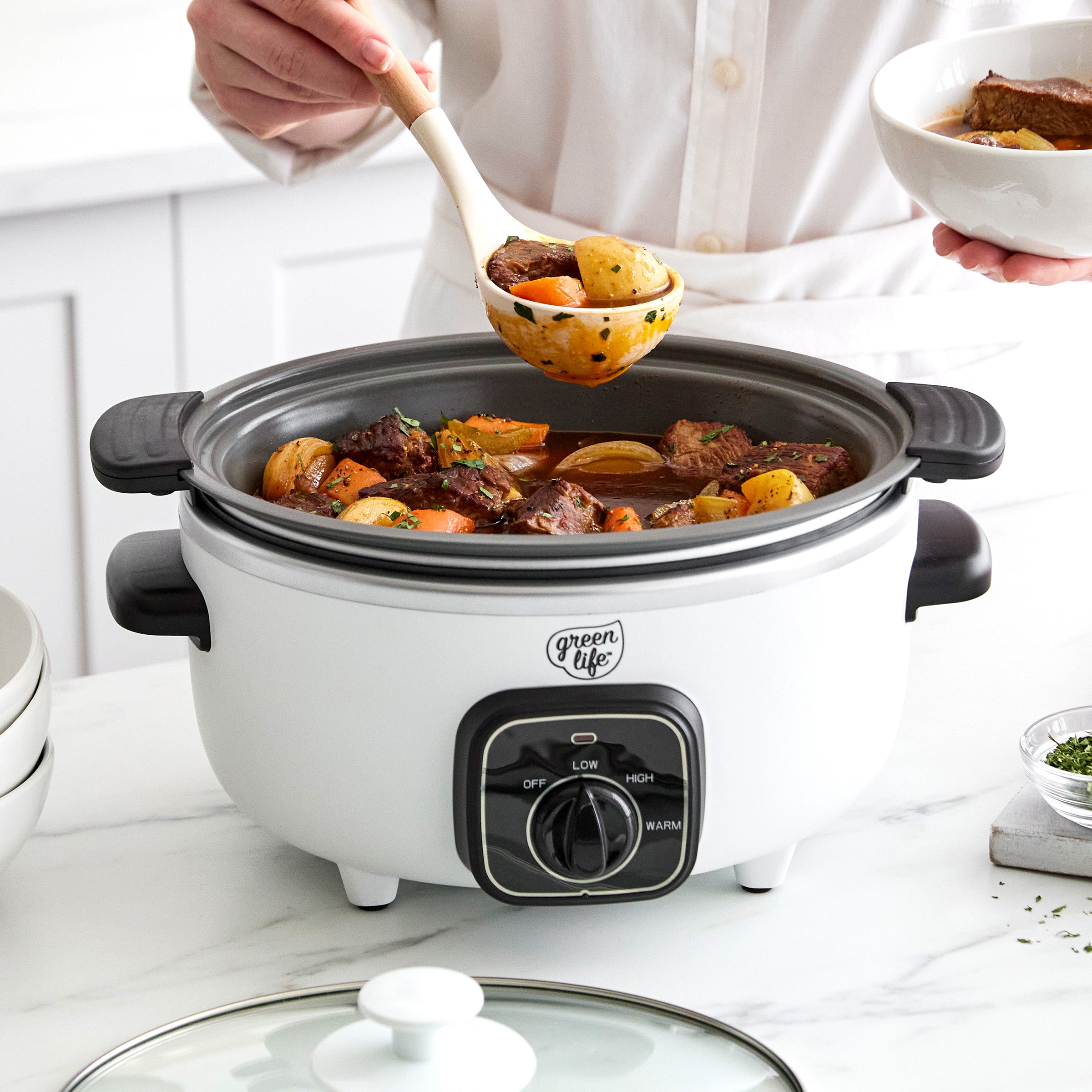 White slow cooker with beef stew, person serving with a ladle, on a countertop