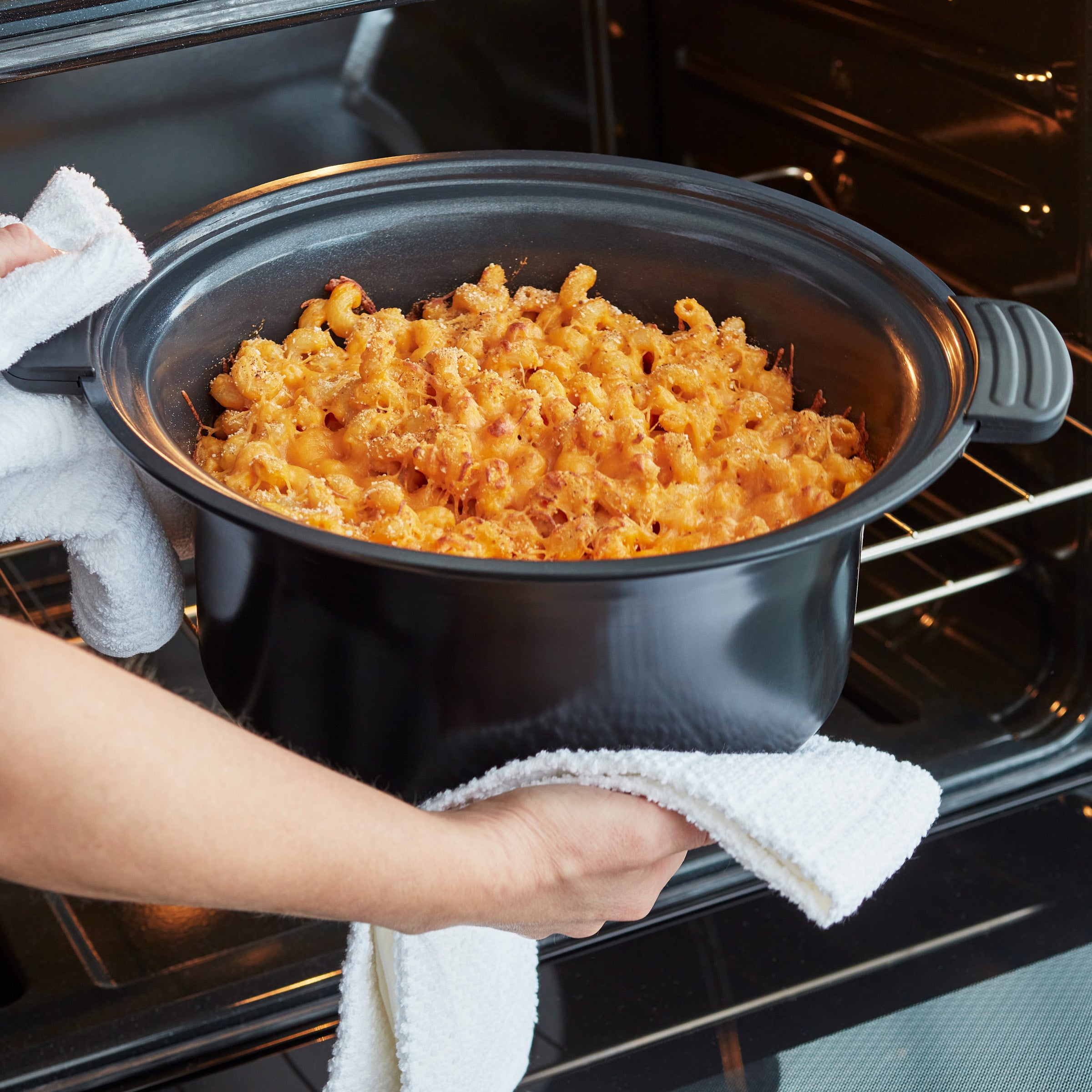 Person removing a dish of macaroni and cheese from an oven using a towel.