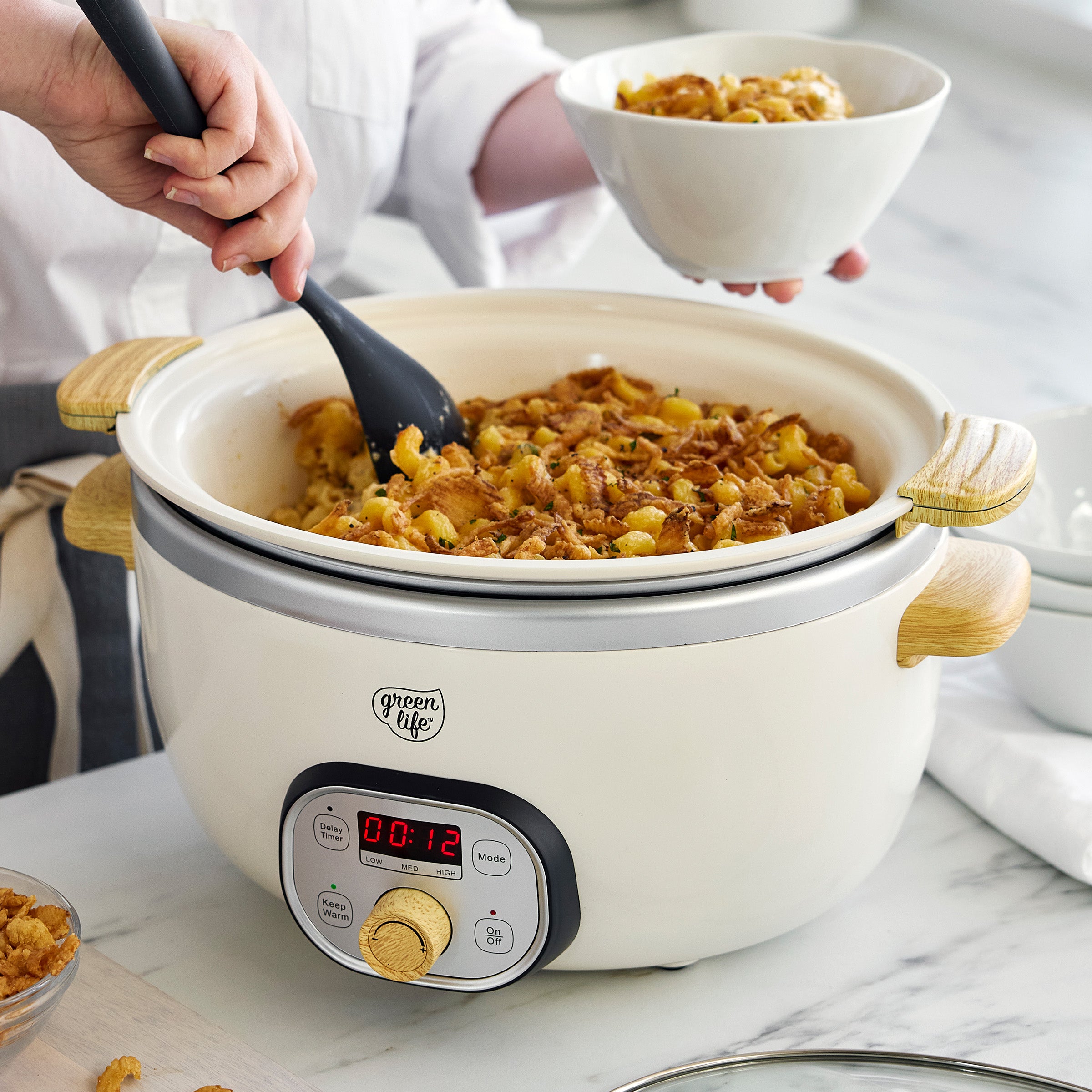Person using a spoon to serve food from a cream slow cooker on a marble countertop.